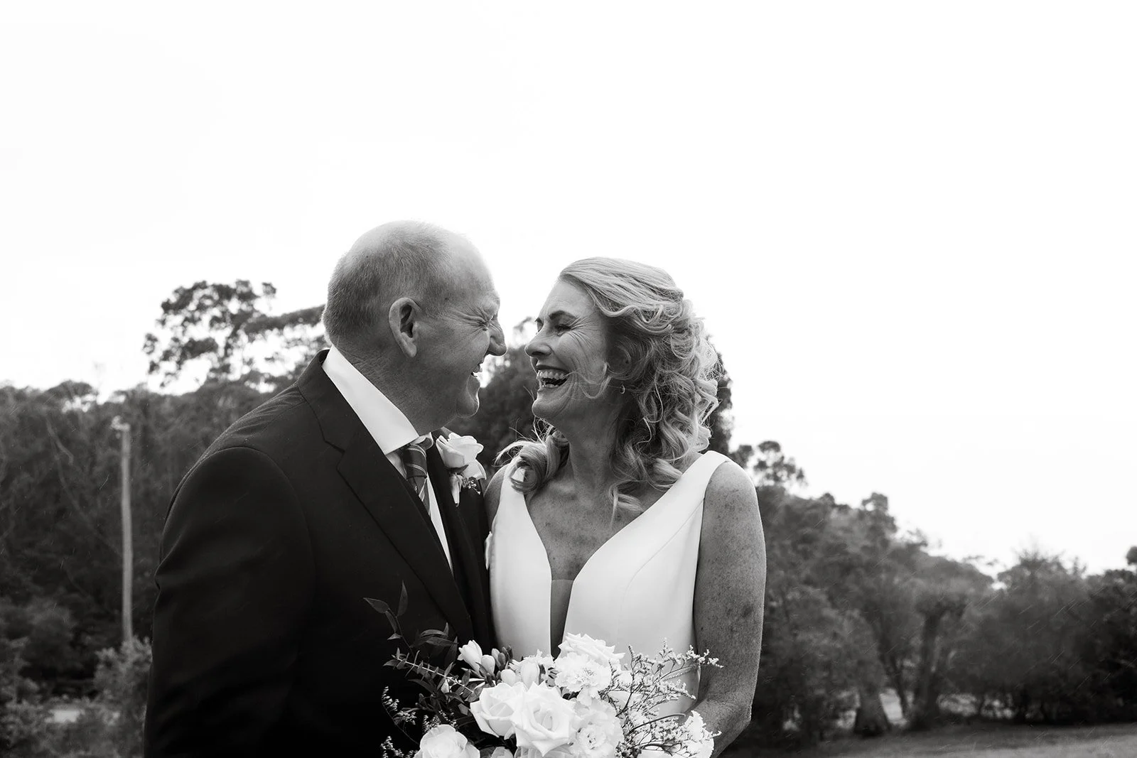 Black and white photo of a joyful middle-aged couple laughing and facing each other outdoors, with a scenic background of trees.