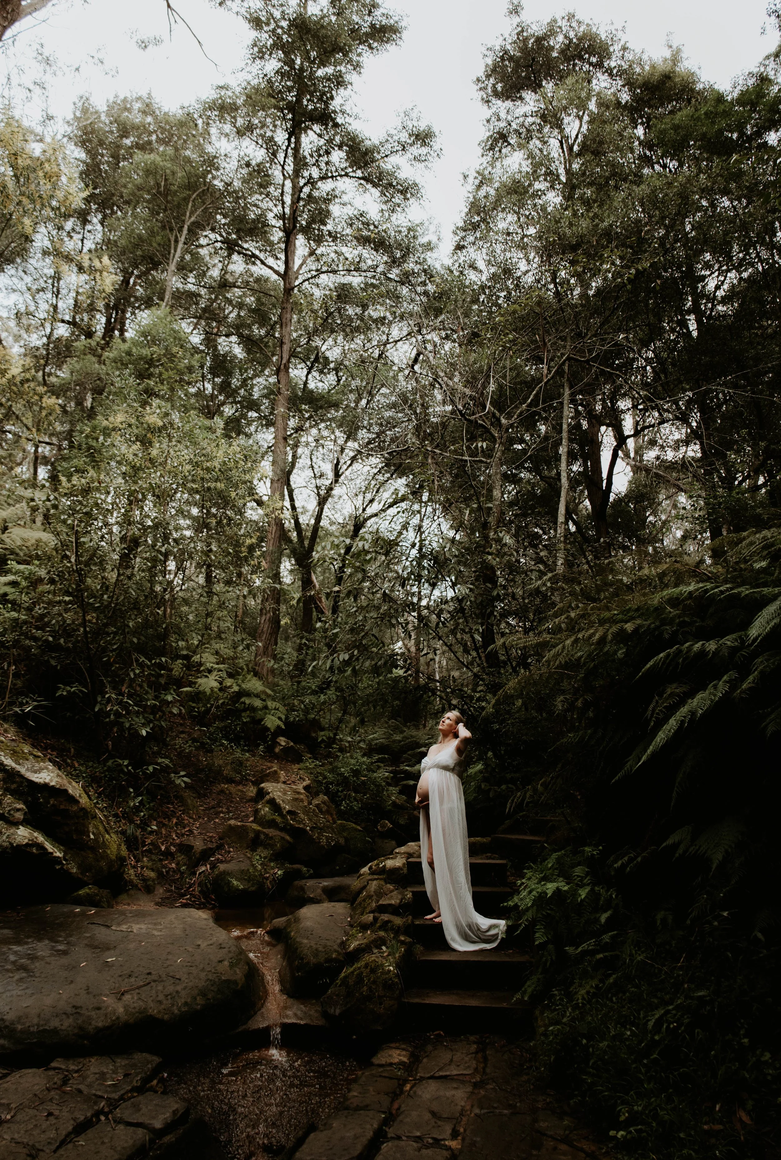 Pregnant woman in a flowing white dress standing on steps in a lush, green forest, looking up with one hand on her head.