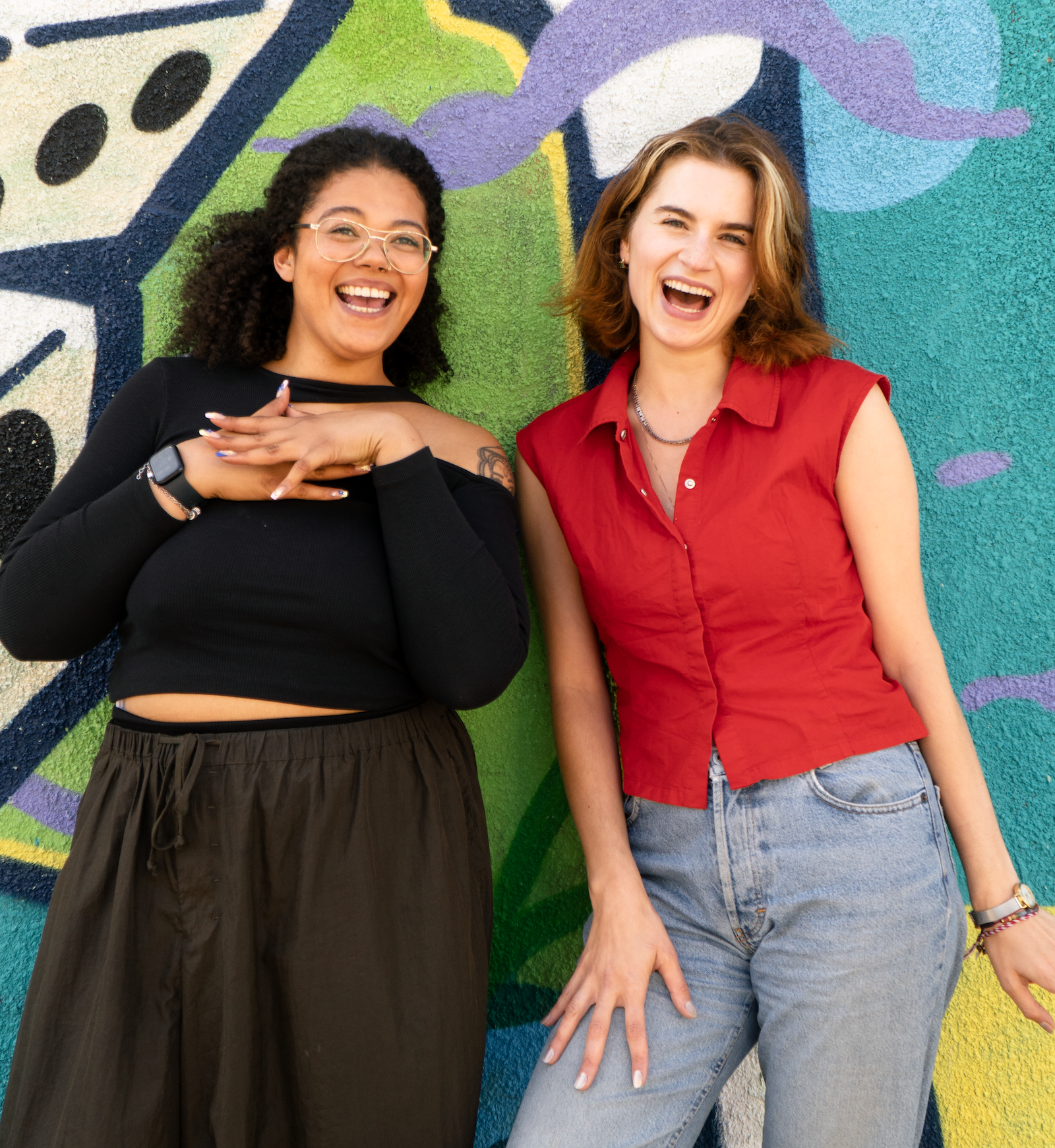 A tall white woman (Mitoshka) in a red top is stood next to a slightly less tall mixed-race woman (Ellayah) with her afro out. They have big smiles on their face and look very happy posting in front of a green graffiti wall in Stokes Croft