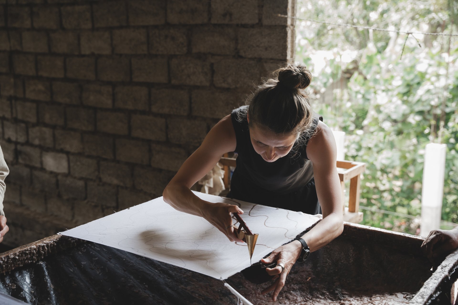 A woman with a bun hairstyle wearing a black sleeveless top is using a tool to carve or work on a large paper or fabric on a table, in a workspace with a brick wall and a window showing green foliage outside.