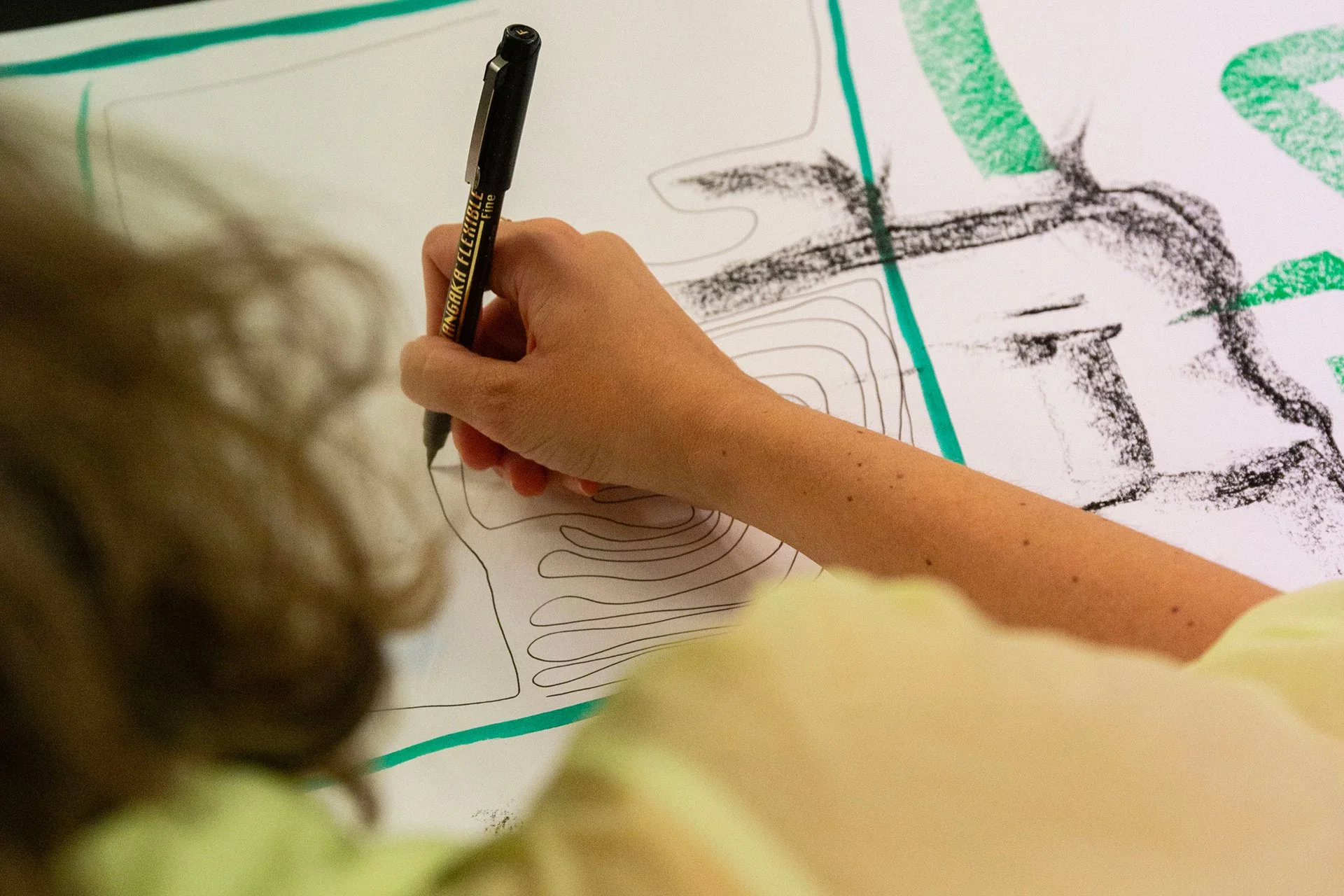 A person is drawing contour lines on a large white sheet of paper with a black marker, with some green and black markings already present on the paper.