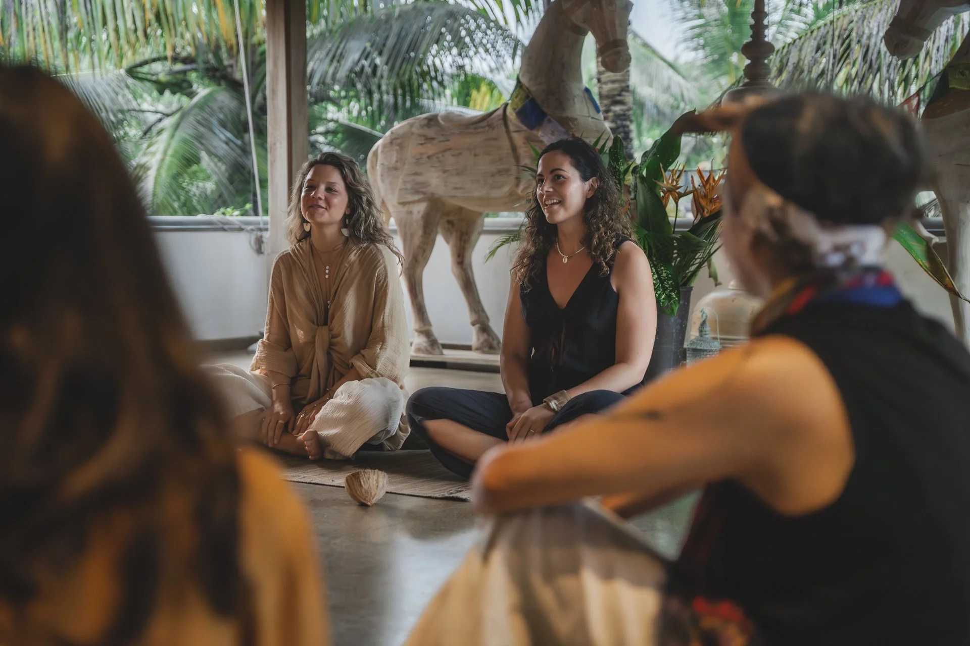 A group of women sitting on the floor in a circle, engaging in a discussion or meditation, with a large wooden horse sculpture and tropical plants in the background.