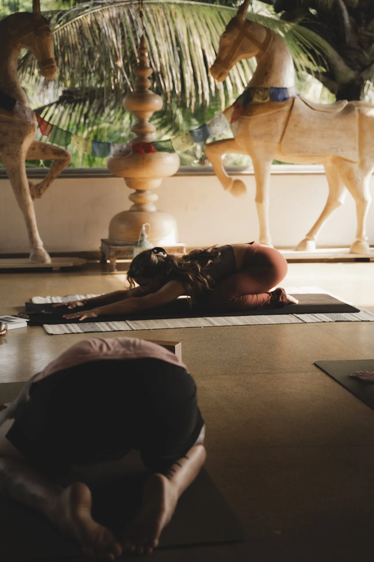 A woman practicing yoga in a studio with large wooden horse and horse statue decorations, tropical plants visible through the window.
