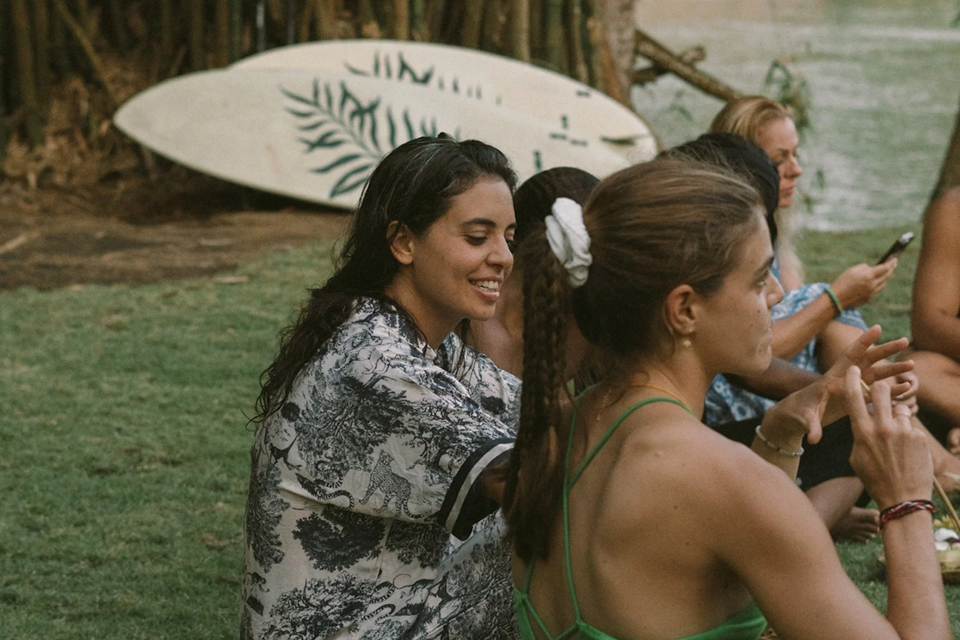 Group of women sitting outdoors on grass near a wooden fence, with surfboards leaning against the fence in the background.