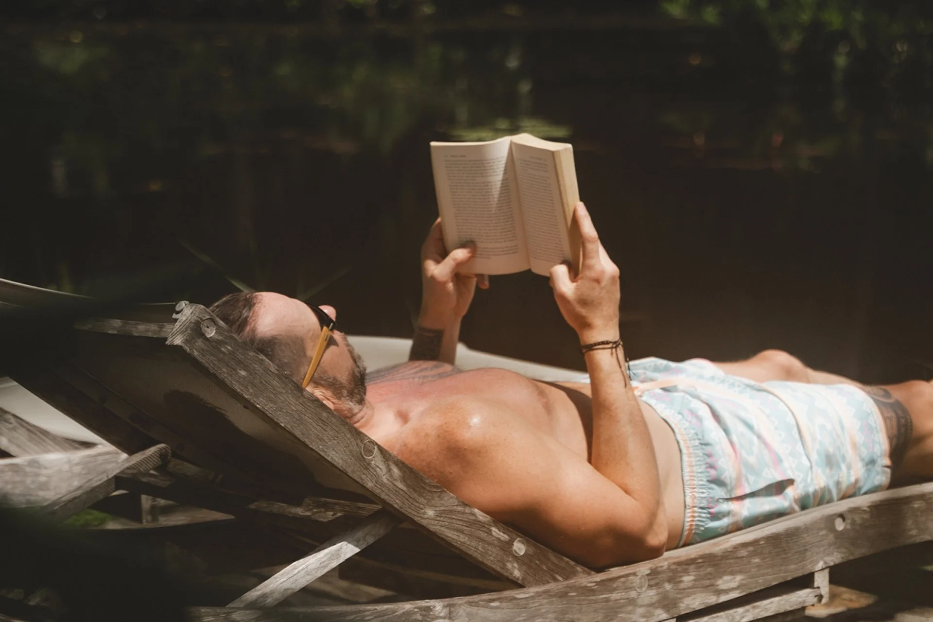 A man lying shirtless on a wooden lounge chair outdoors, reading a book, with a forest background.