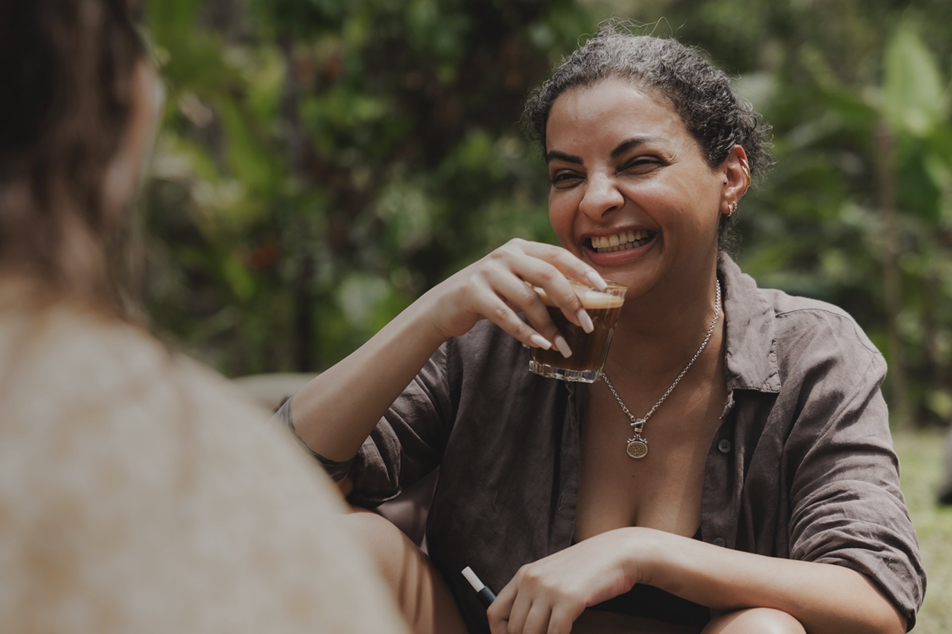 Two women sitting outdoors, one smiling and holding a shot glass of dark beverage, the other with back turned, in a lush green environment.
