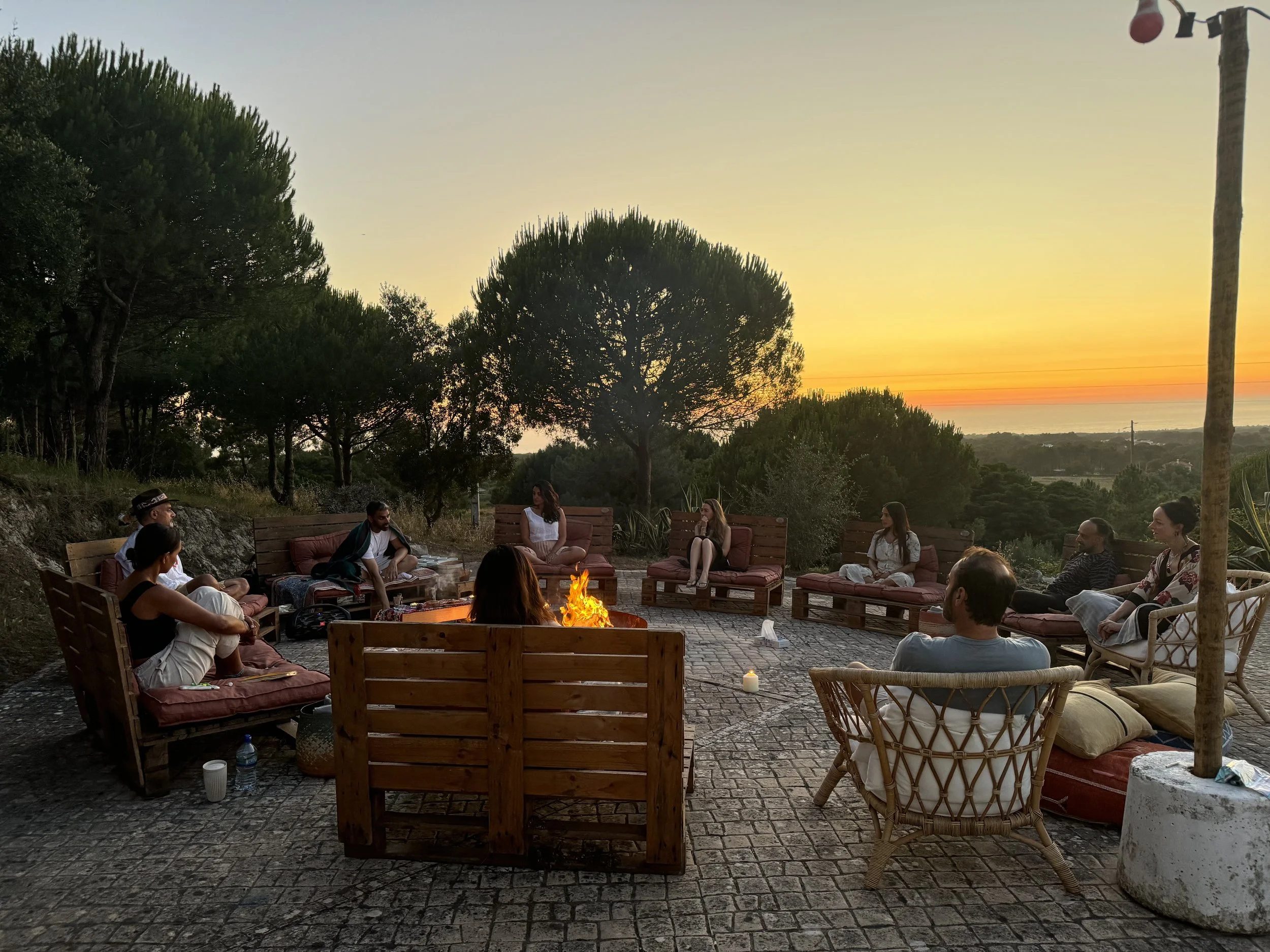 Group of people sitting around a fire pit outdoors at sunset, surrounded by trees and scenic view.