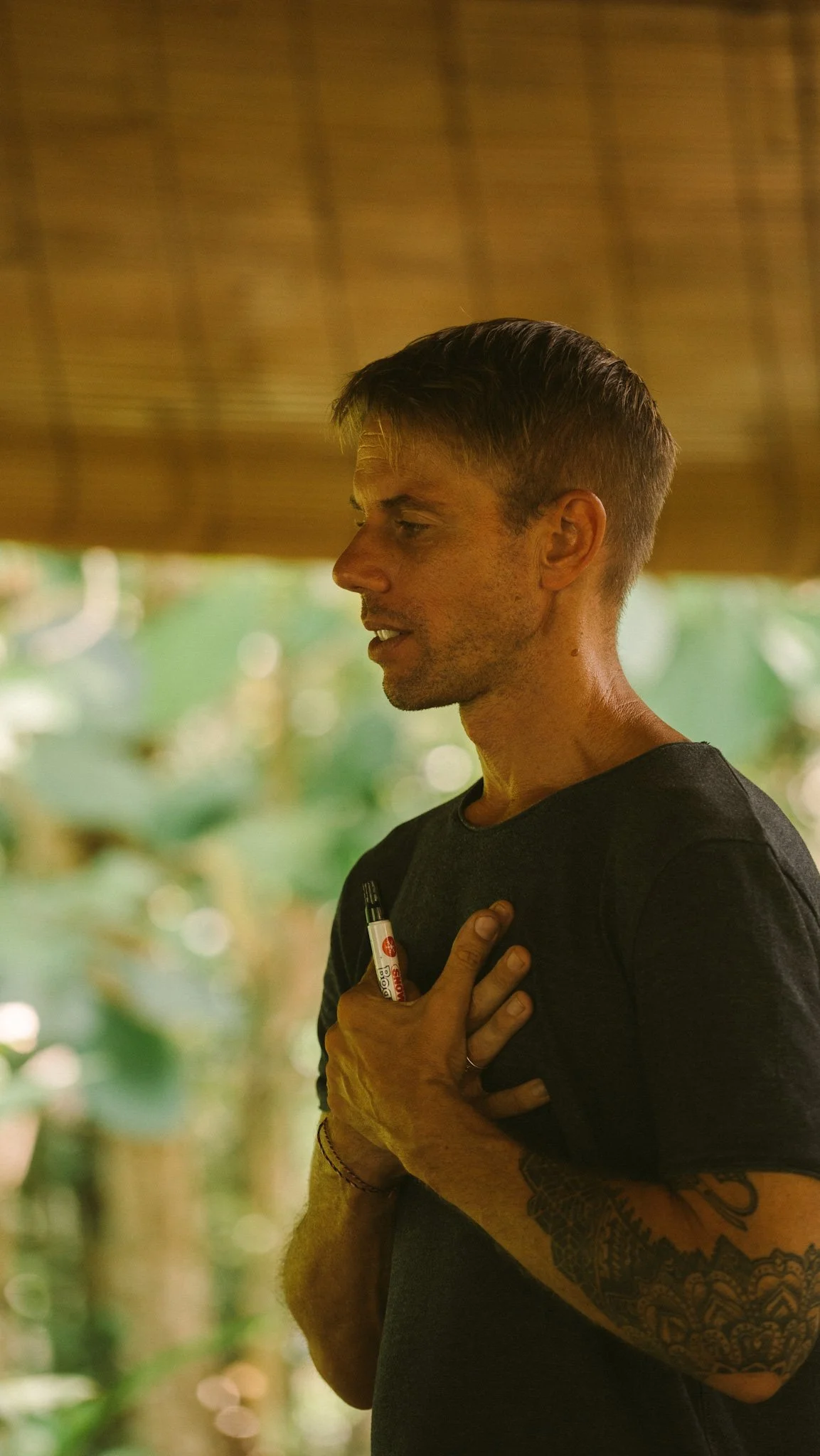 A man with short hair and tattoos on his arm is holding a marker in his pocket and placing his right hand on his chest, appearing to be praying or expressing gratitude, in a lush, green, outdoor setting.