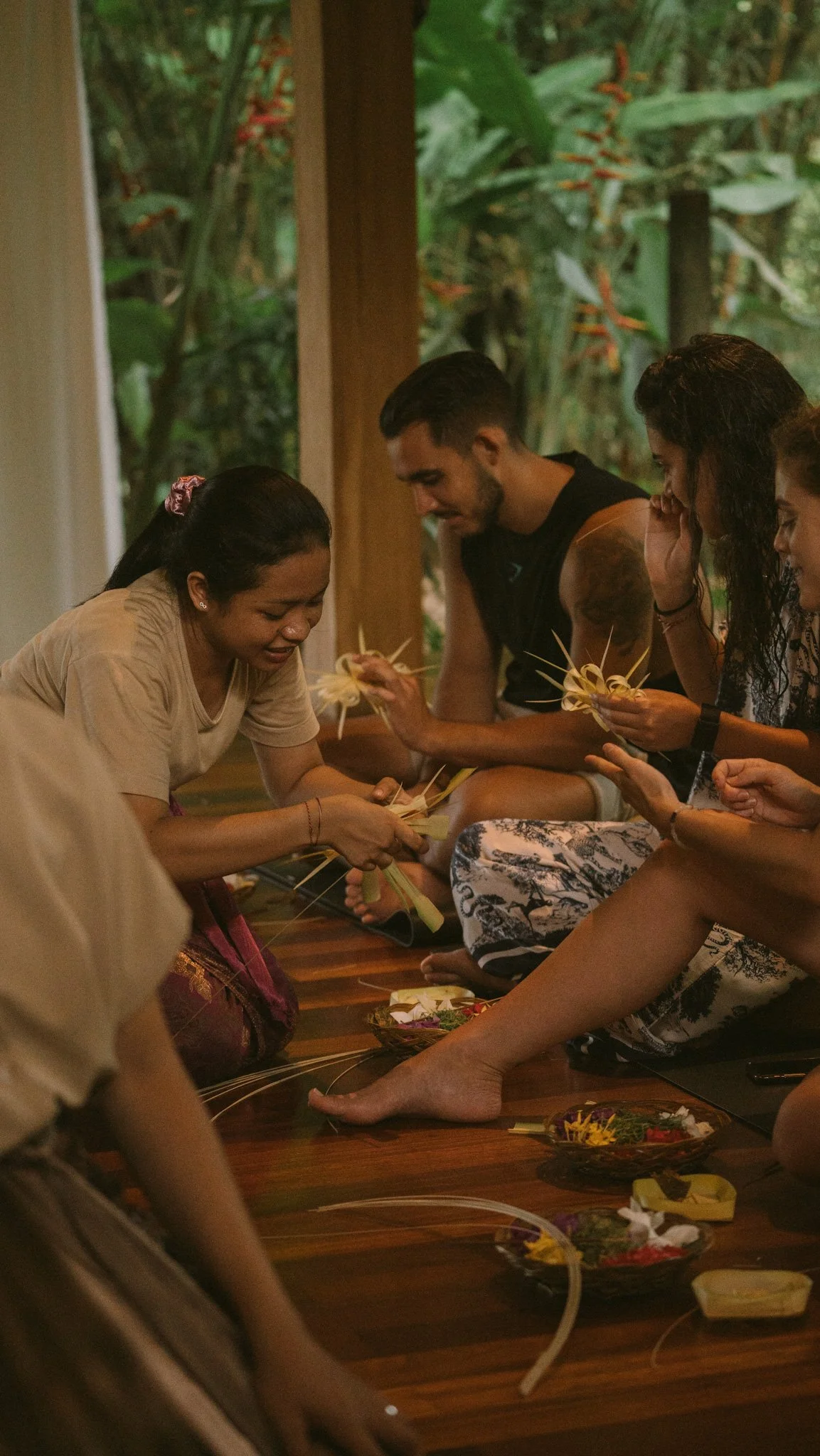 People sitting on the floor, making traditional woven crafts with flowers and leaves, inside a room with large windows and lush greenery outside.