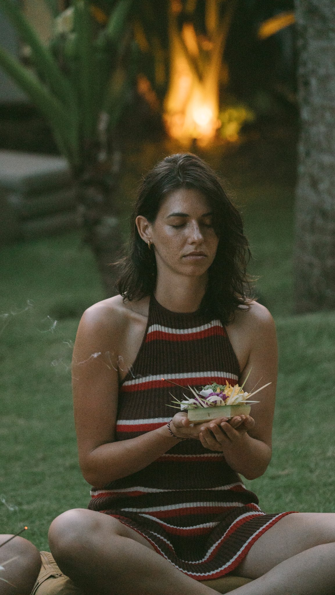 A woman with dark hair and medium skin sits cross-legged on the grass during dusk, holding a small dish with flowers, with green plants and warm lighting in the background.
