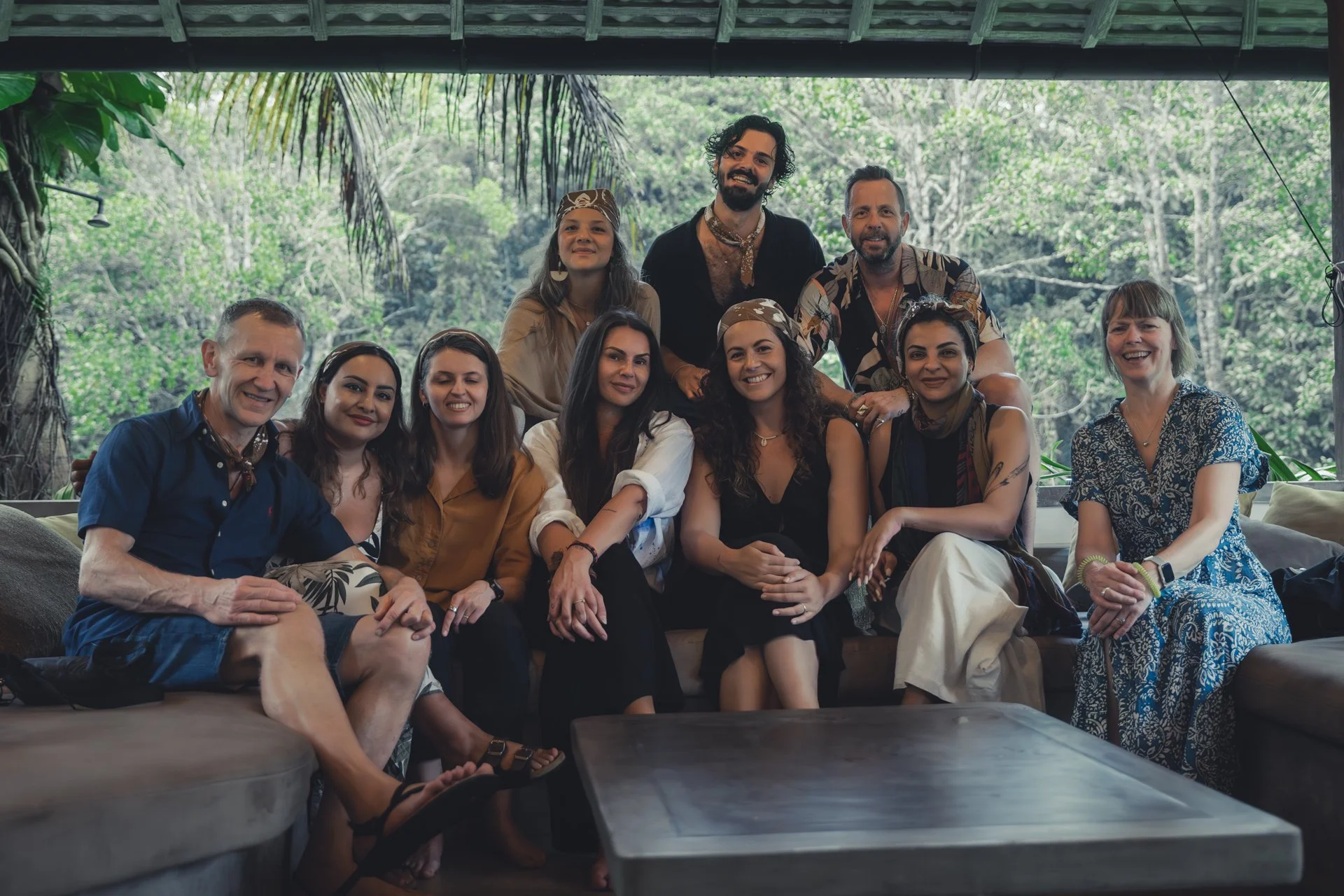 Group of ten diverse people smiling and posing for a photo in a tropical outdoor setting with lush green trees in the background.