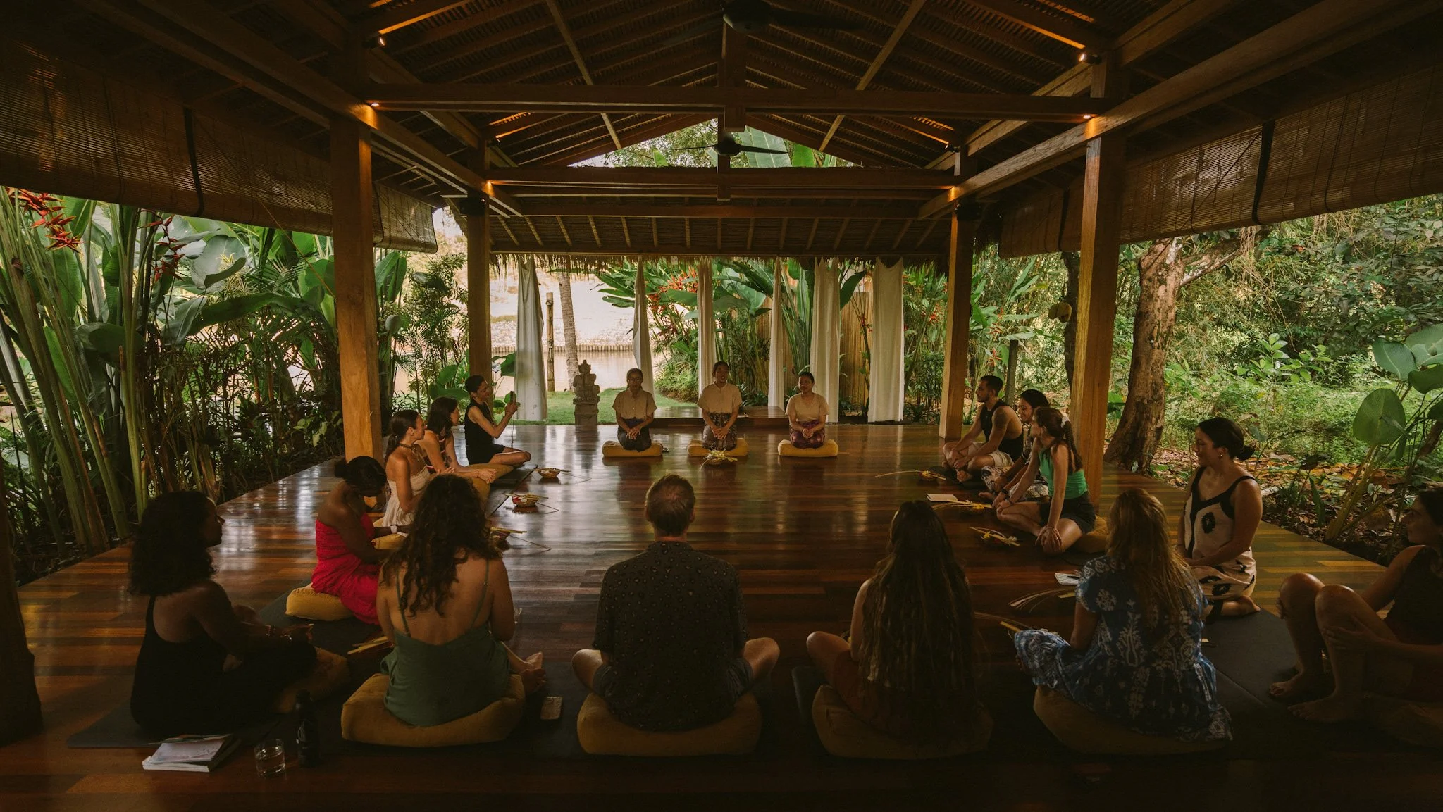 People participating in a meditation or yoga session in a lush, open-air wooden pavilion surrounded by greenery.