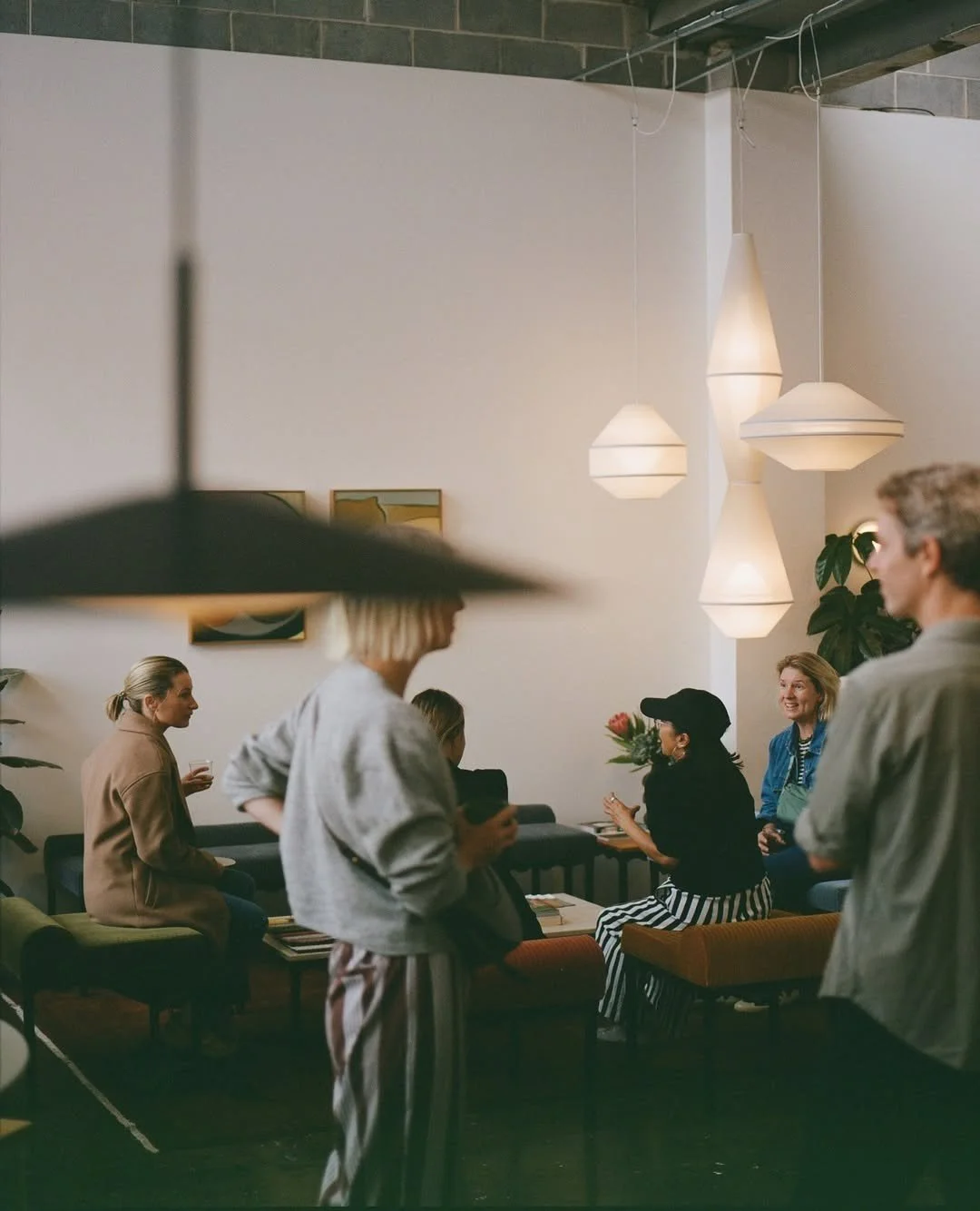 Group of people talking and socializing in a modern, well-lit indoor space with hanging pendant lights, art on the walls, and a cozy seating area.
