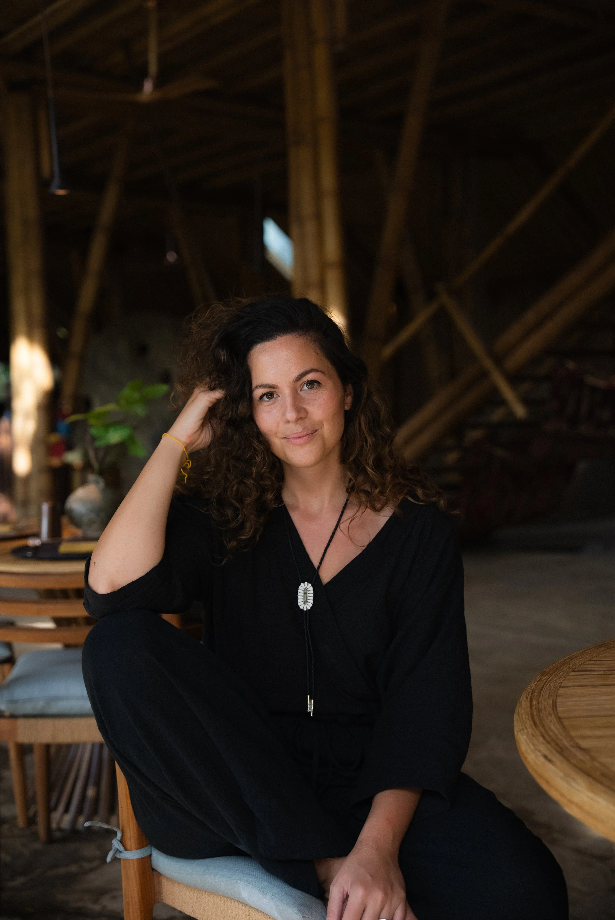A woman with curly brown hair and tan skin sitting on a chair inside a rustic wooden structure. She is wearing black clothing and a necklace, and is smiling slightly while looking at the camera.