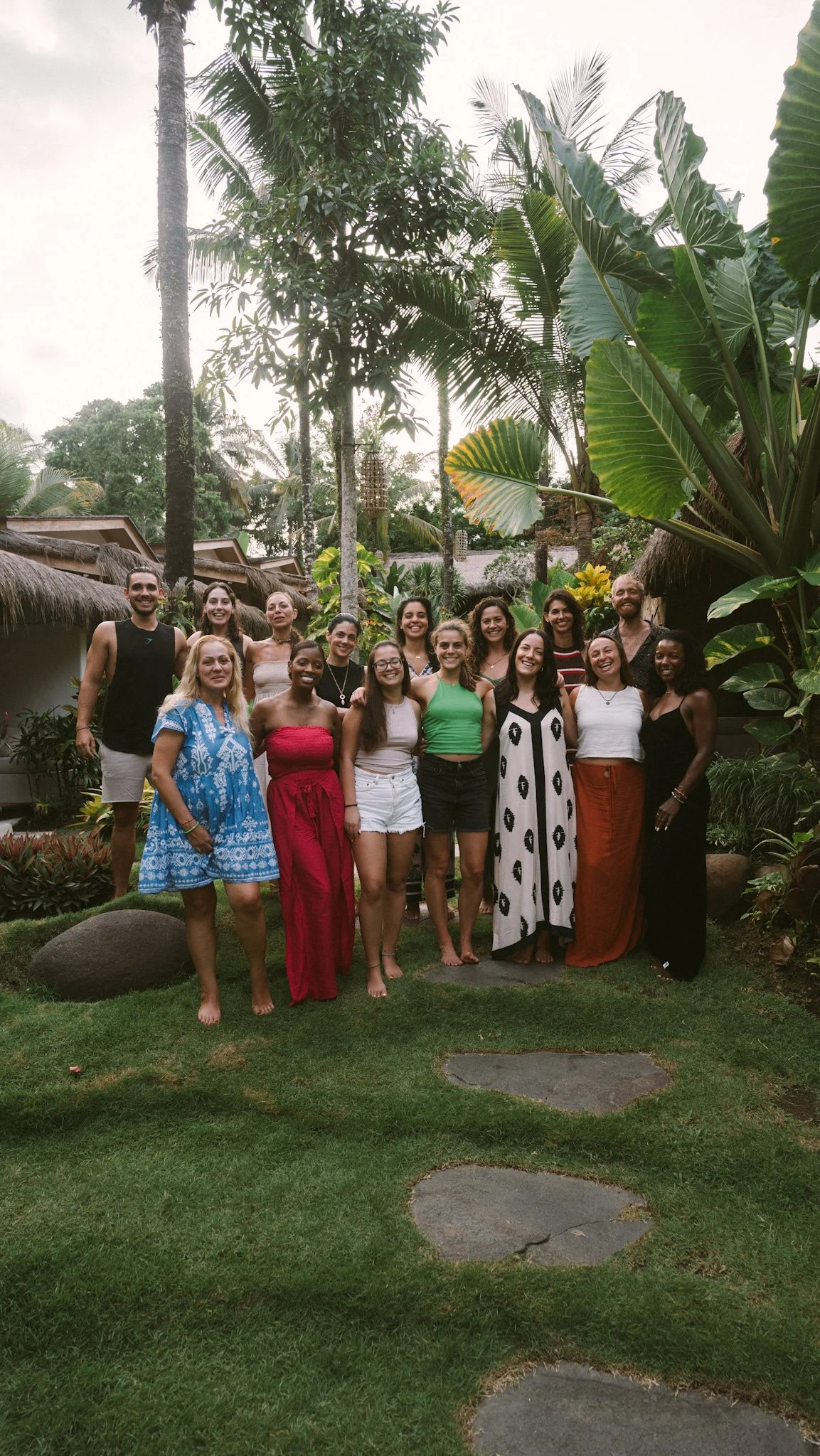 A group of fifteen people standing together outdoors in a lush tropical garden, smiling for the photo.