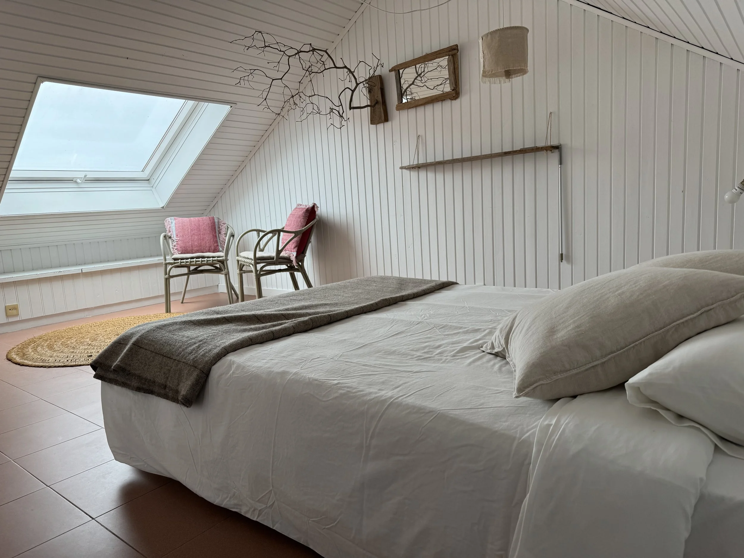 A bedroom with a white bed and pillows, a large skylight window, two chairs with pink cushions, a circular rug, and decorative wall items including a mirror, branch, and shelving.
