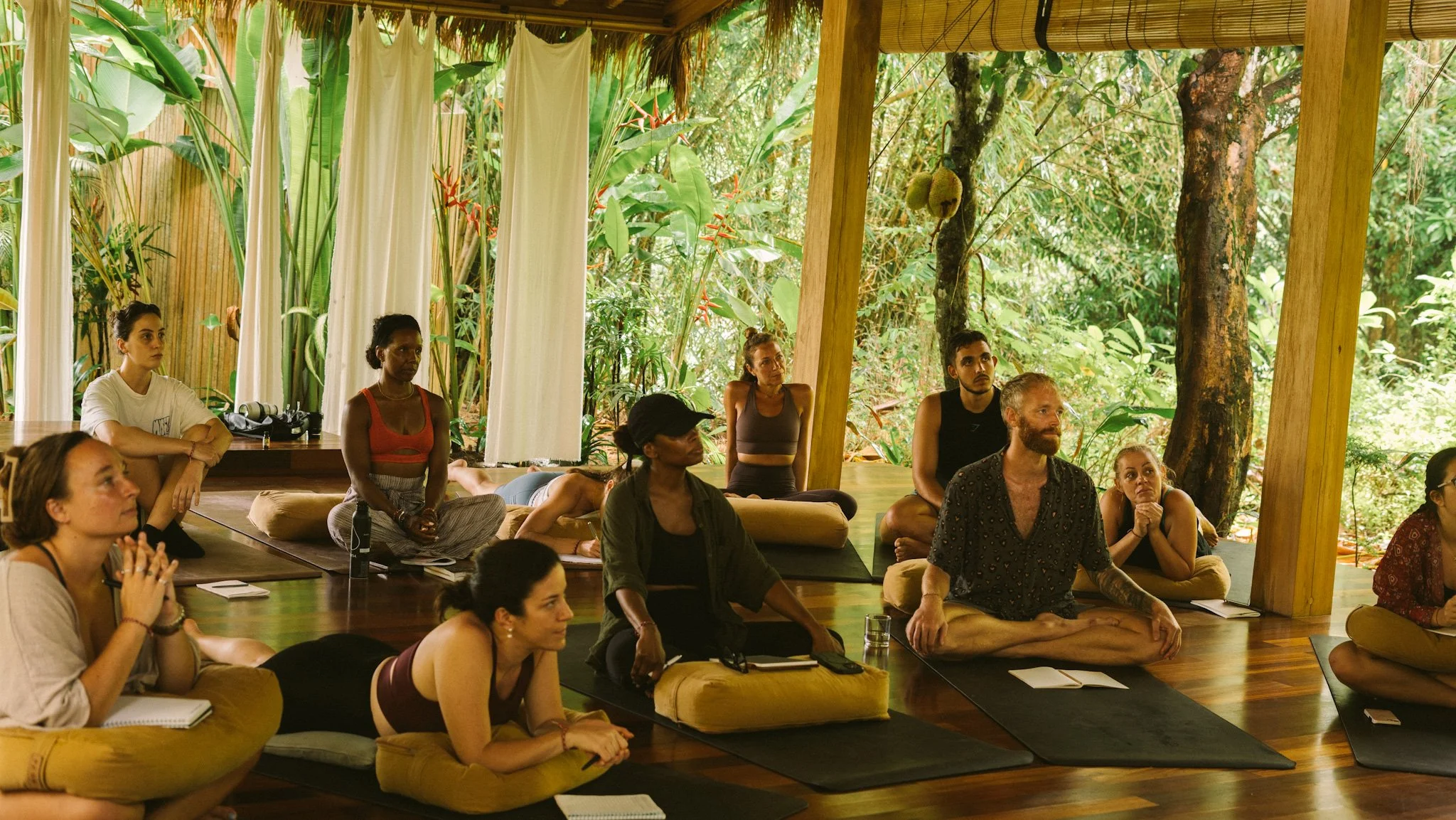 People practicing yoga or meditation on cushions and mats in a lush, green outdoor wooden pavilion.