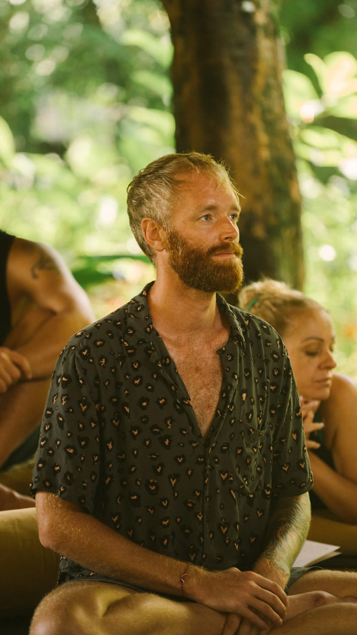 A man with a beard and mustache, wearing a black shirt with a small heart pattern, sitting cross-legged in a lush, green outdoor jungle setting, with a woman beside him, and large trees and greenery in the background.
