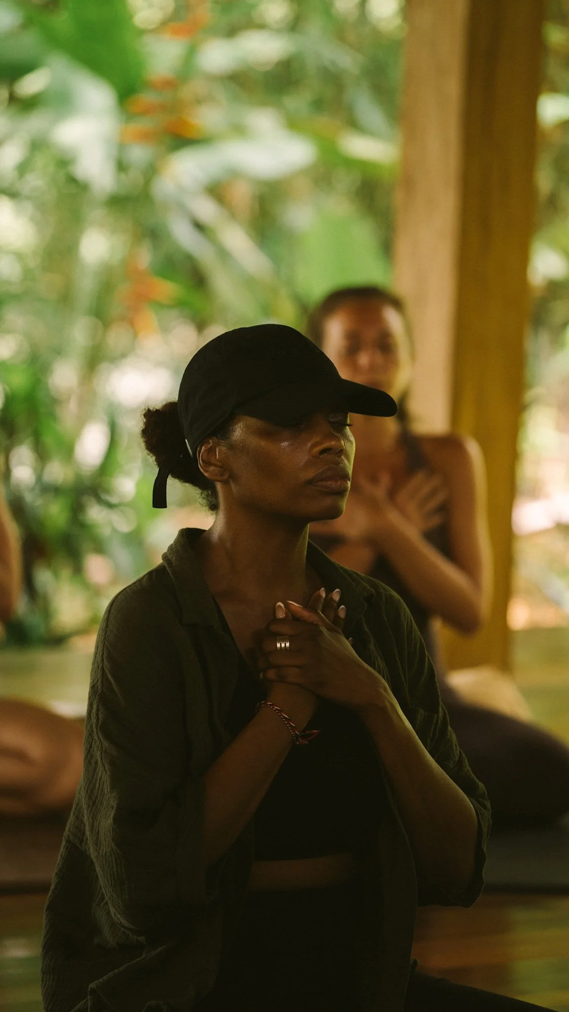 A woman with dark hair pulled back, wearing a black cap and dark clothing, with her hands clasped to her chest and eyes closed. Another woman is blurred in the background, sitting in a natural, green environment.