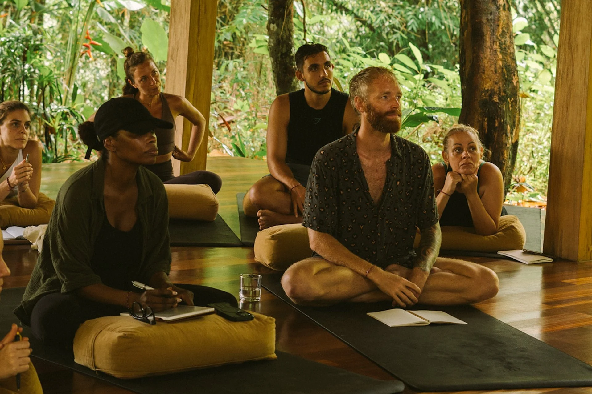 A group of people sitting on cushions and mats on a wooden floor in a lush, green, outdoor setting, engaged in a meditation or yoga session.