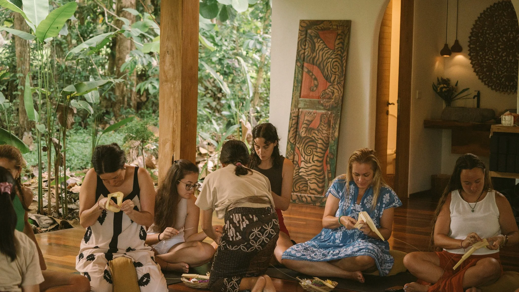 Group of women sitting cross-legged on a wooden floor in a room with large windows overlooking a lush green jungle, engaged in a cultural or educational activity, with traditional artwork and decor on the walls.