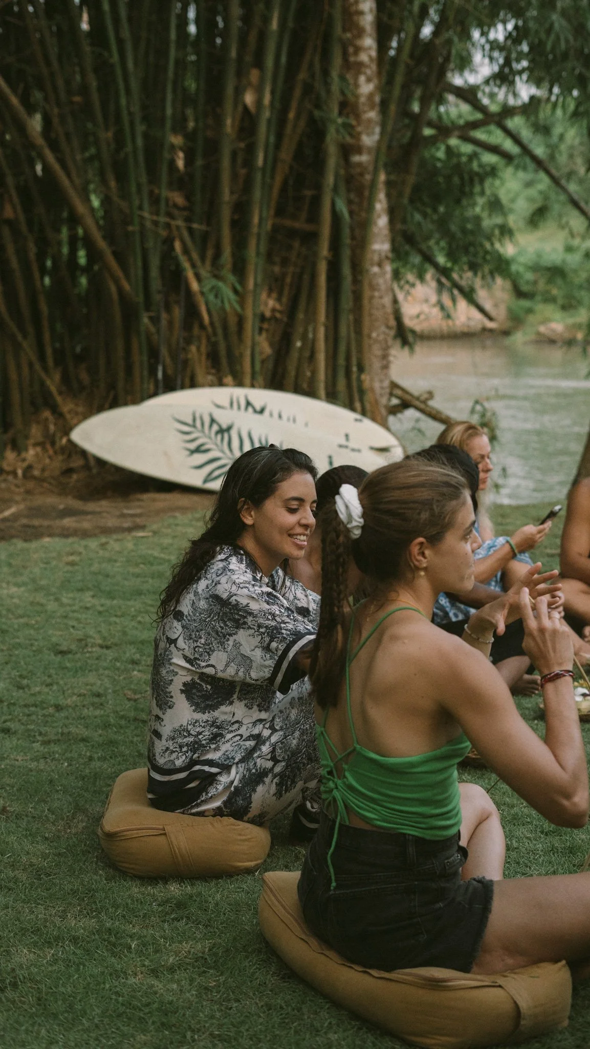 Group of women sitting on cushions by a river in a lush, tropical setting, with a surfboard leaning against trees in the background.