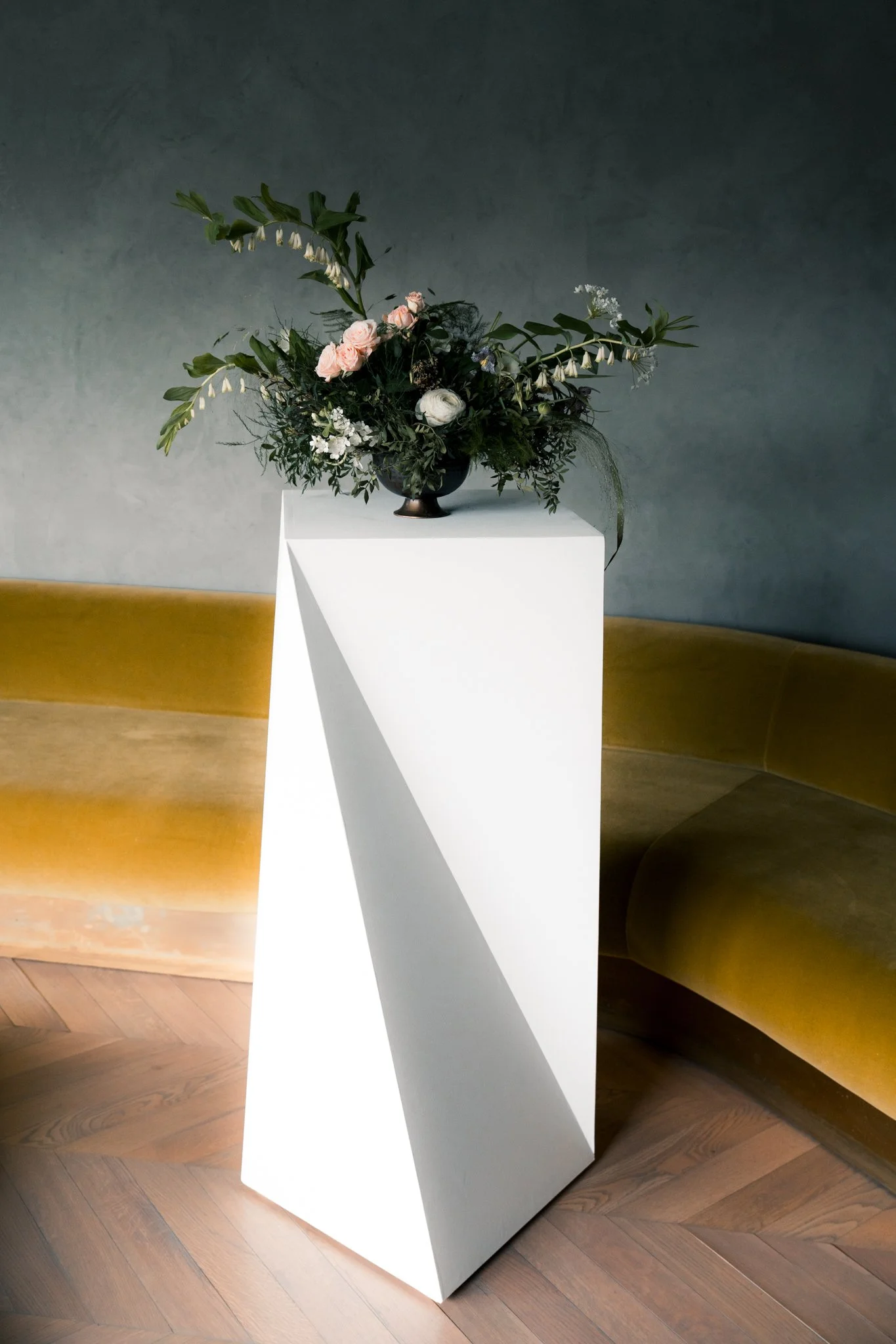 A modern flower arrangement in a black vase on a white pedestal, with a curved gold sofa in the background.