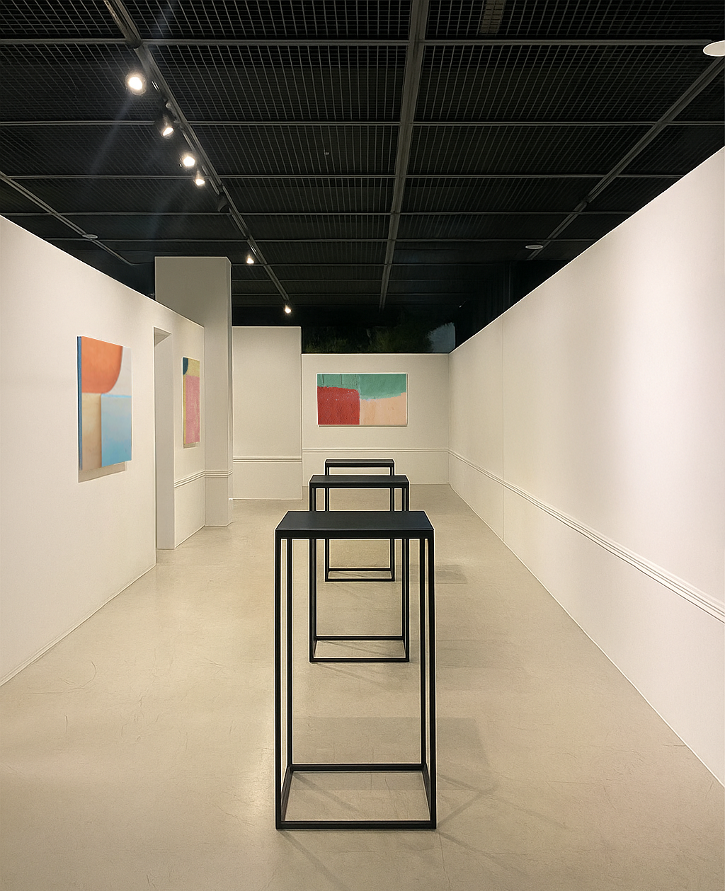 Empty art gallery with four abstract paintings on white walls and three black stools arranged in a row on the beige floor, under a black ceiling with ceiling lights.