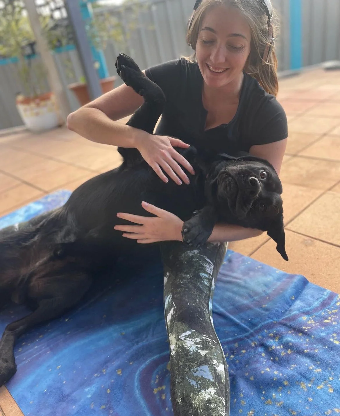 A woman smiling while sitting on a blanket, holding a black dog that is leaning against her. The woman appears to be petting the dog on the chest and belly. The setting appears to be a patio with potted plants and a fence in the background.