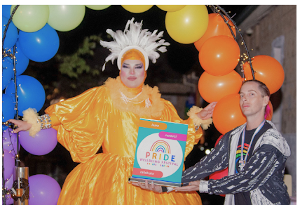 Image from previous Busselton Pride Wellbeing Festival events featuring a Drag Queen performer in an orange gown and white headress.