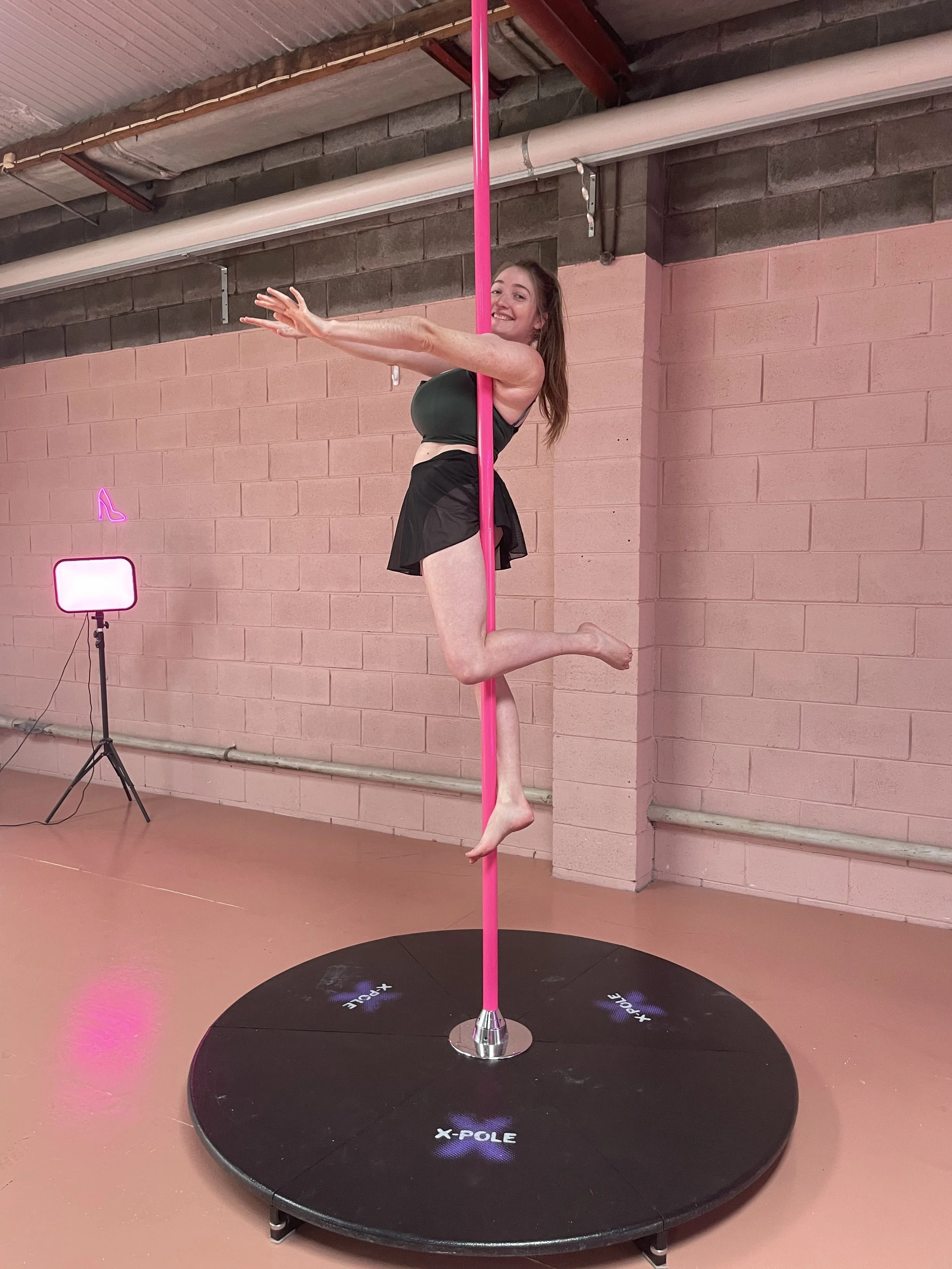A woman performing pole dance on a pink pole in a studio with pink and gray walls and pink neon sign.