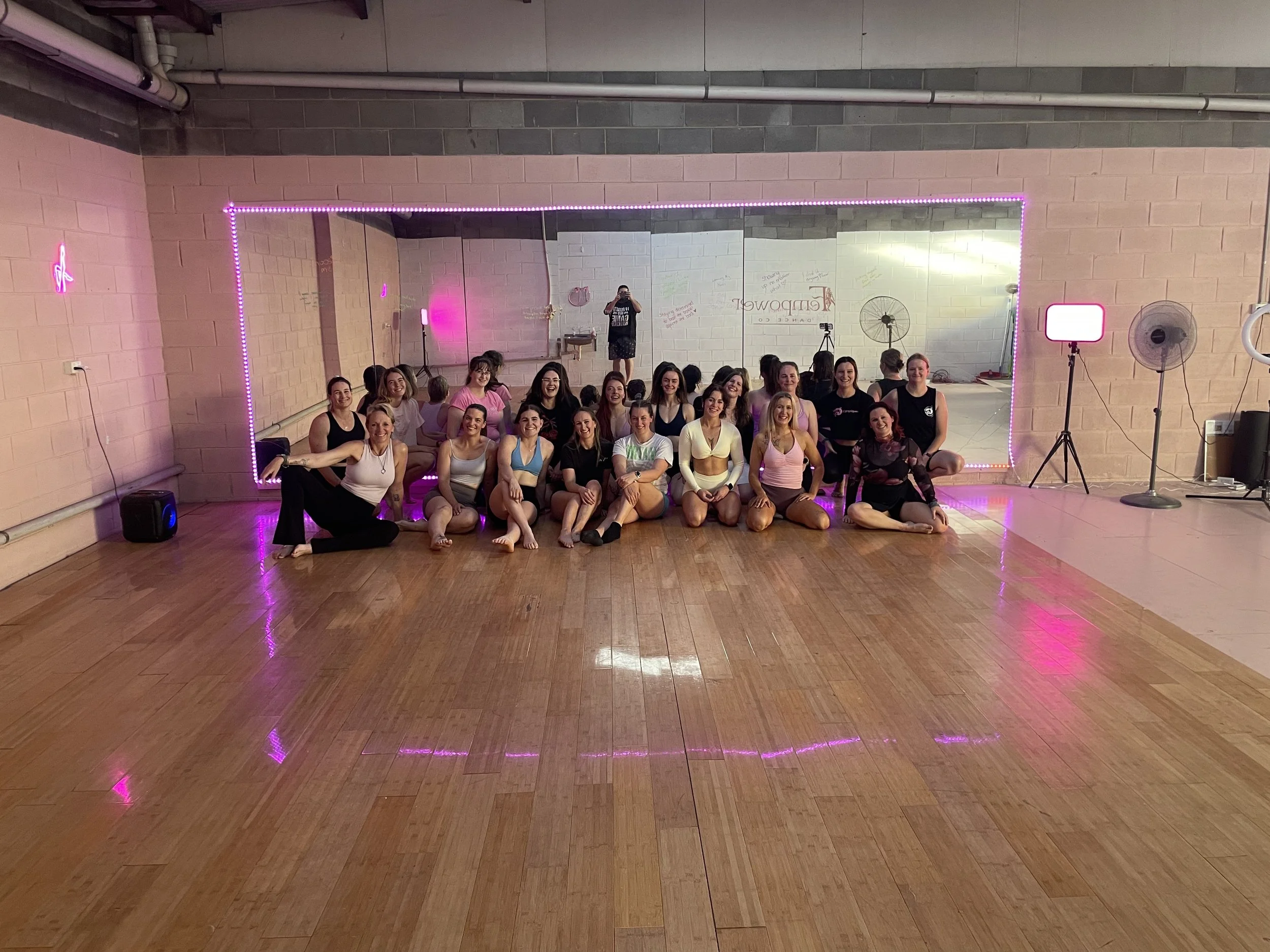 Group of women in fitness attire posing for a photo in front of a mirror with purple LED lights at a dance studio.