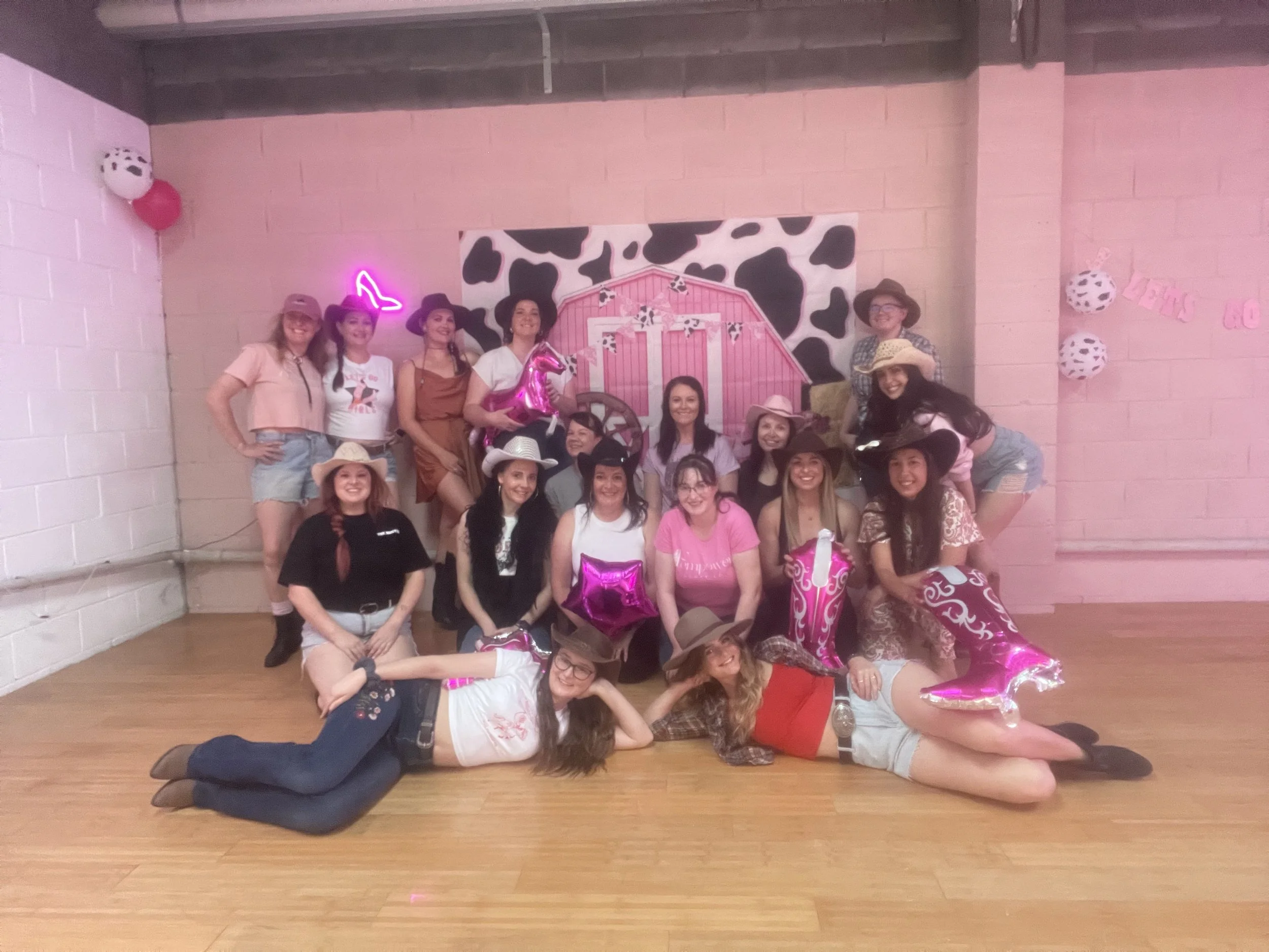 A group of mostly young women dressed in cowgirl and cowboy costumes, some wearing hats, gathered in a dance studio with pink decor and cow print background, celebrating in a Western-themed party.
