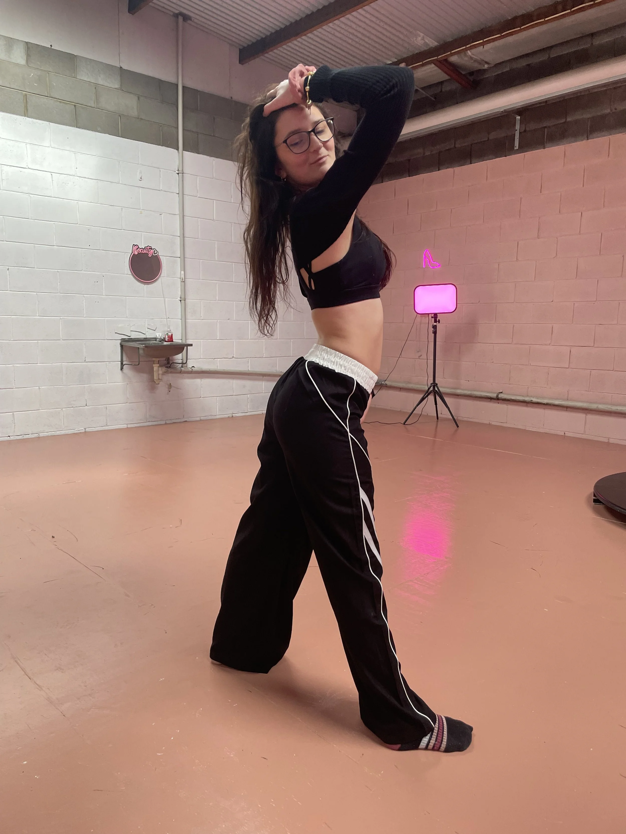 A young woman with glasses and long hair is in a dance studio or rehearsal space with exposed brick and cinderblock walls, and pink neon light in the background. She is wearing a black crop top, black track pants with white stripes, and socks. She is posing with one hand on her forehead and eyes closed, smiling faintly.