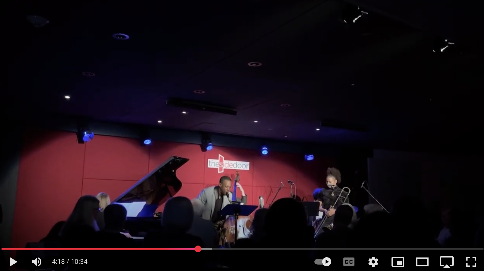 Jazz musicians performing on stage at The Side Door, including a pianist, a double bass player, and a trombone player, in front of a red backdrop.