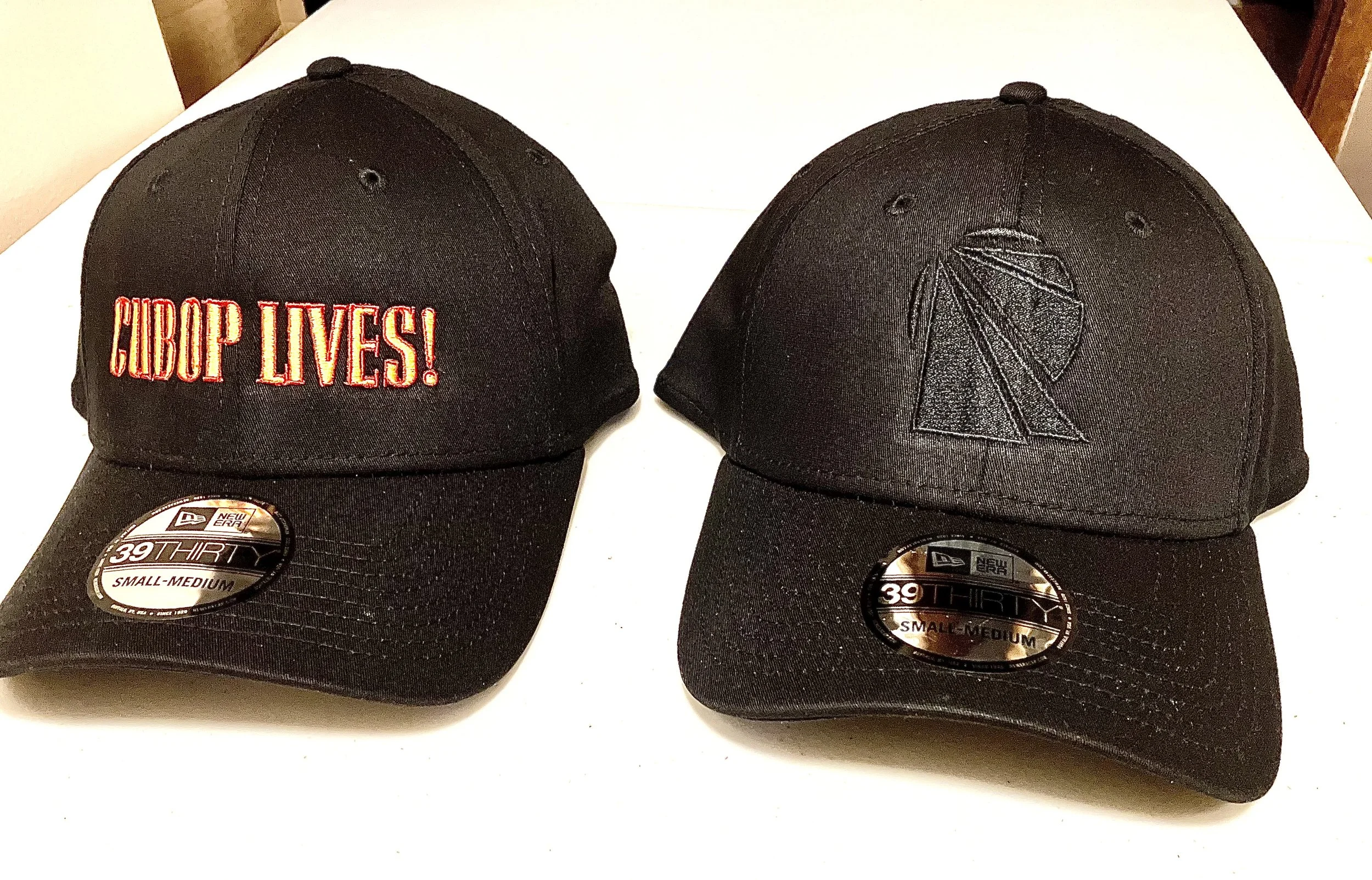 Two black baseball caps, one with red embroidered text reading 'Gibop Lives!' and the other with black embroidery of a marijuana leaf. Both caps have stickers indicating they are size small-medium. They are placed on a white surface.