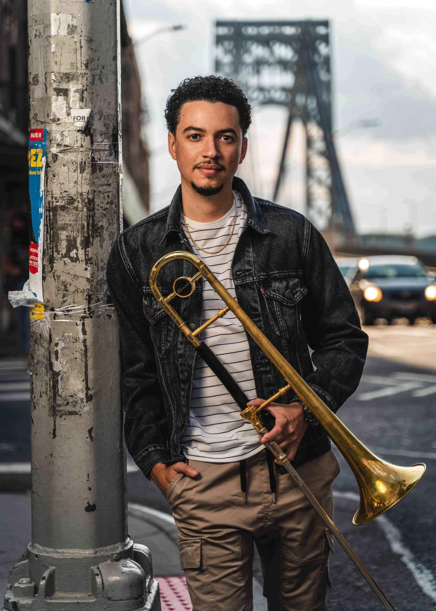 A man in a beige hoodie and black pants sitting on a stool in a recording studio with a brass instrument, headphones, and microphone.