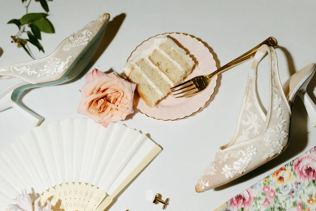 A slice of white cake with layers and icing on a pink plate, a gold fork resting on the plate, a pink rose beside the plate, high-heeled shoes with lace and embroidery, a white paper fan, and a floral fabric pattern.