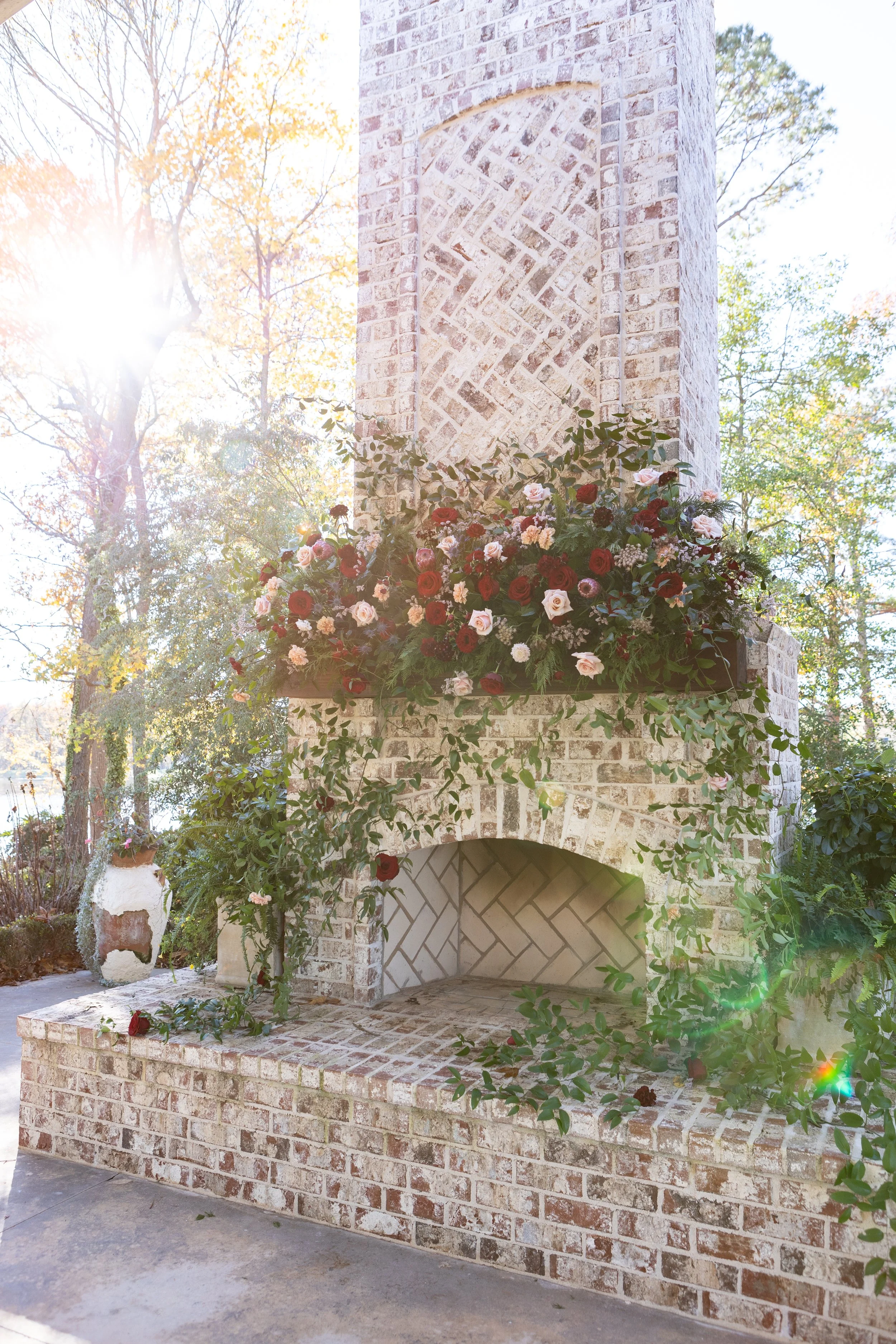 Brick outdoor fireplace decorated with a large floral arrangement of red, pink, and white flowers, with greenery, on a sunny day.