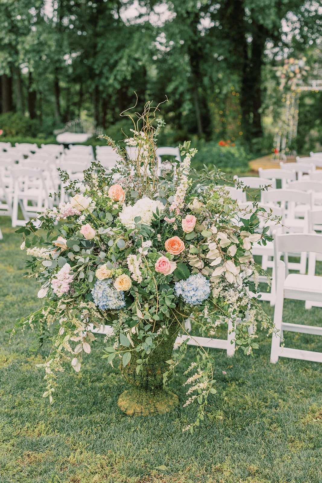 A large floral arrangement with roses, hydrangeas, and greenery in front of white chairs at an outdoor wedding ceremony.