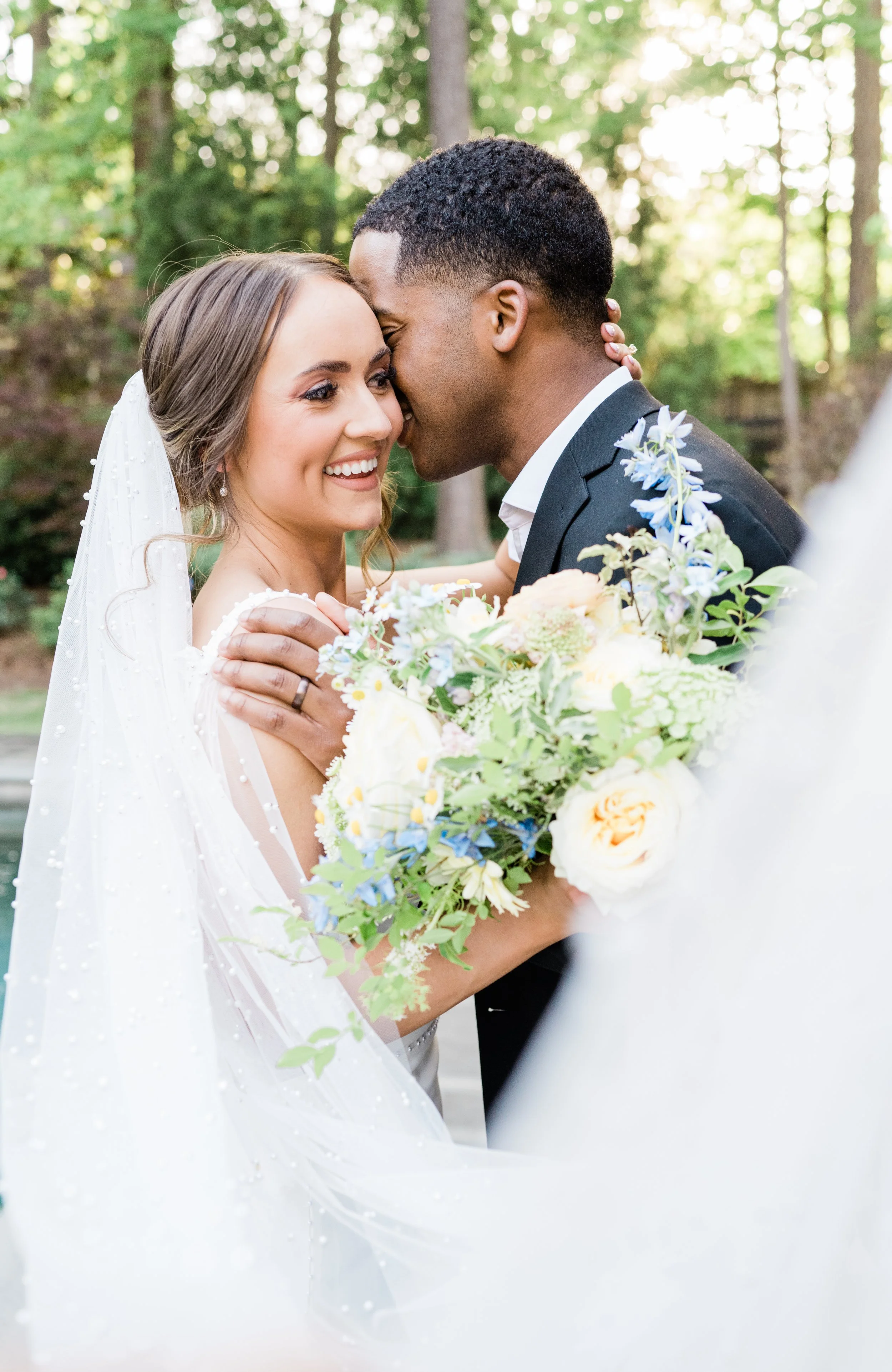 A bride and groom sharing a close, intimate moment outdoors, with the bride holding a bouquet of flowers and smiling, surrounded by lush greenery.