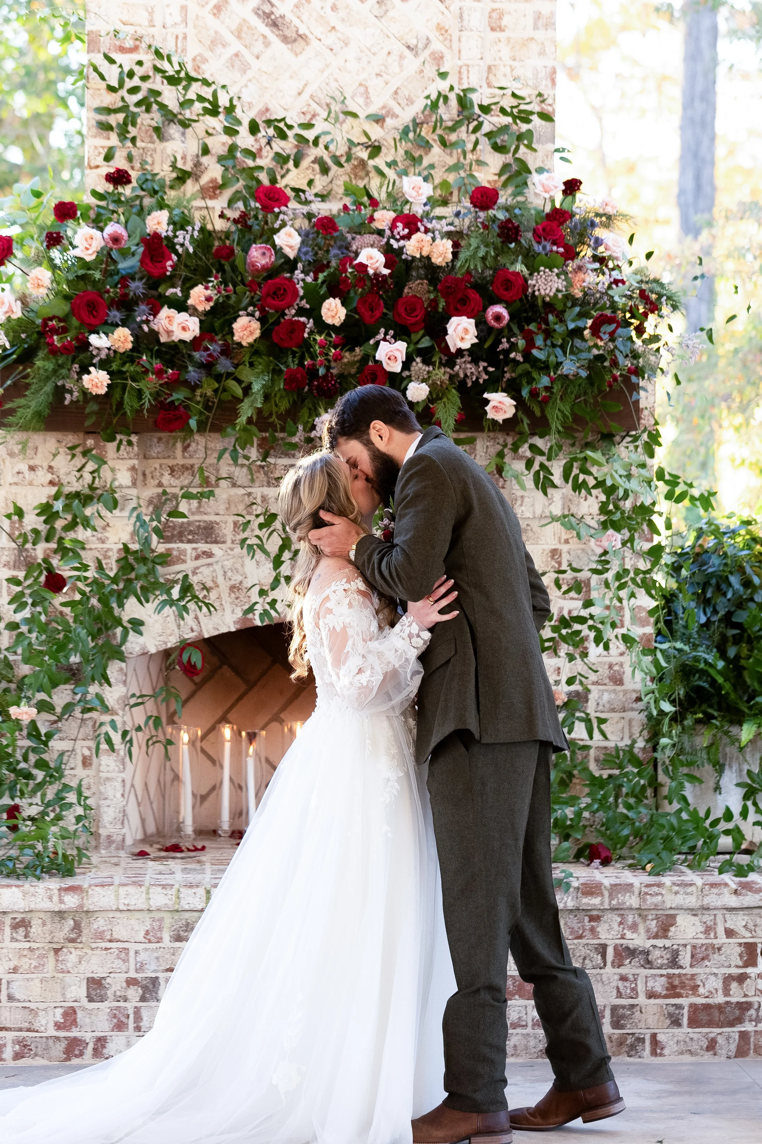 A bride and groom sharing a kiss during their wedding ceremony in front of a brick fireplace decorated with a large arrangement of red, pink, and white flowers and greenery.