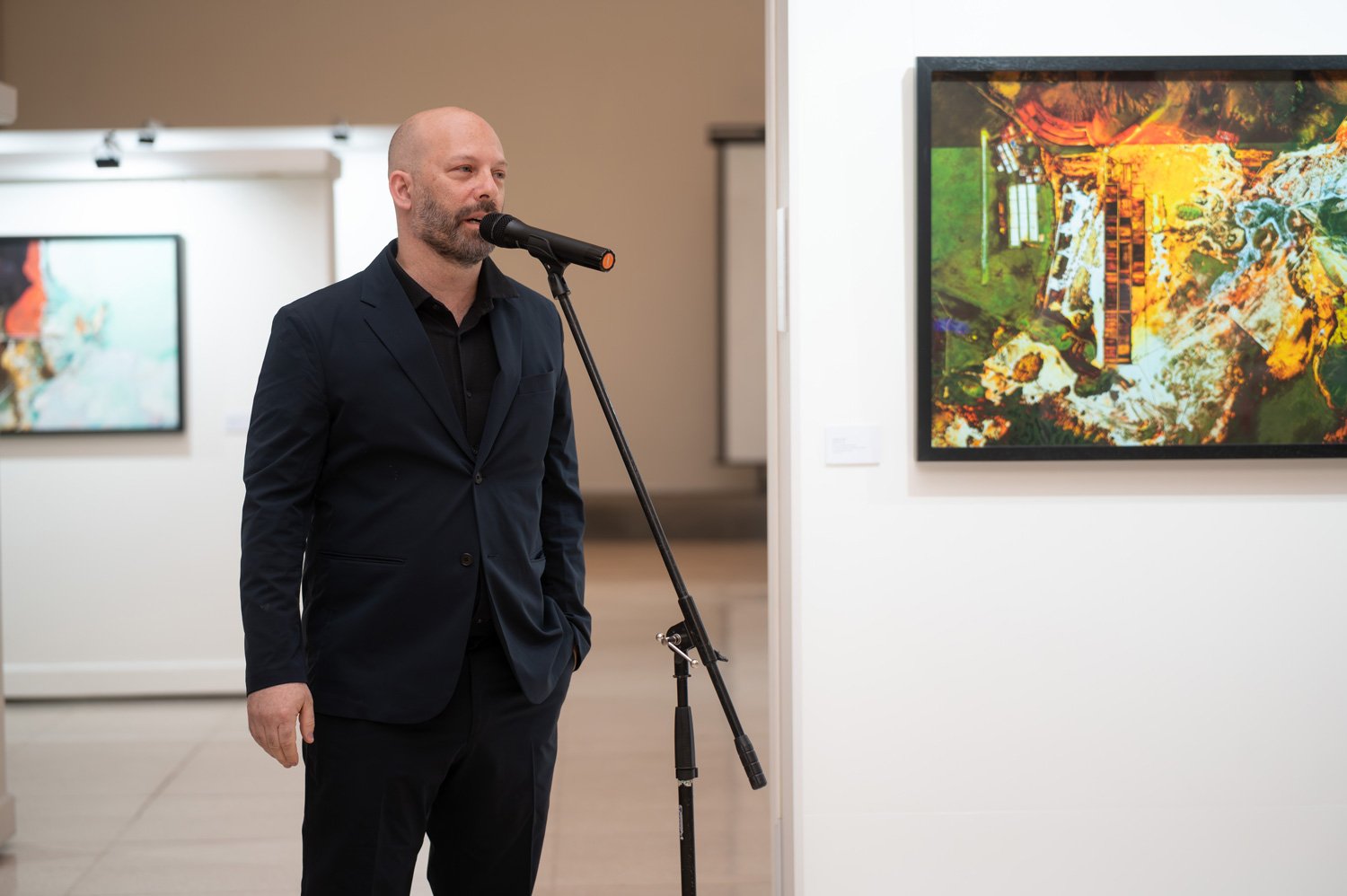 A man in a black suit and black shirt speaks into a microphone at an art gallery. Artwork is displayed on white walls behind him.