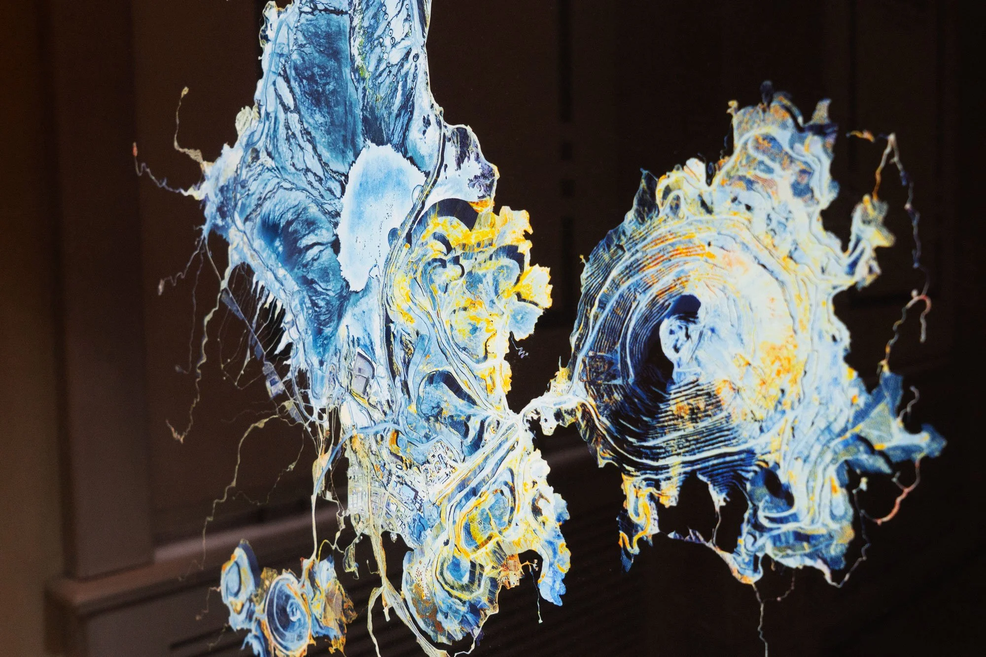 Abstract artwork resembling geographical maps with intricate blue, yellow, and white patterns on a dark background.