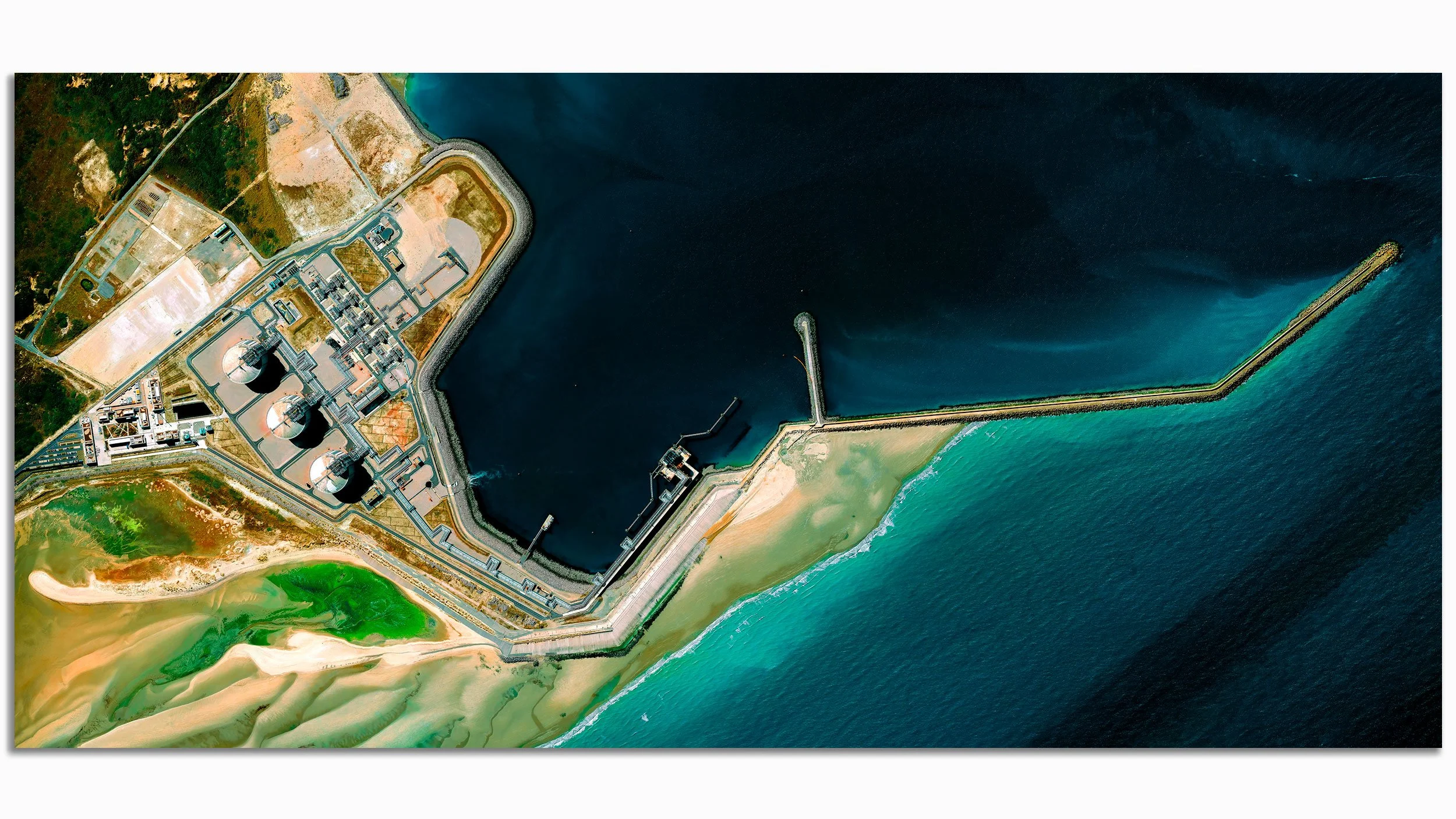 An aerial view of a large water treatment facility with tanks, structures, and a sea wall, located by the ocean with sea and sand beaches.
