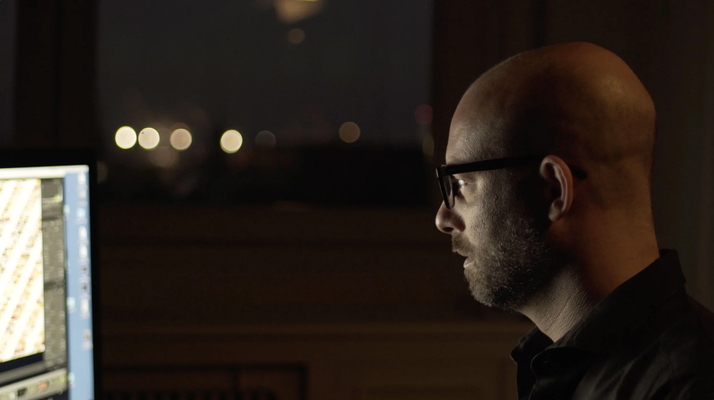 A man with glasses working on a computer at night, with a dark background and blurred city lights outside the window.