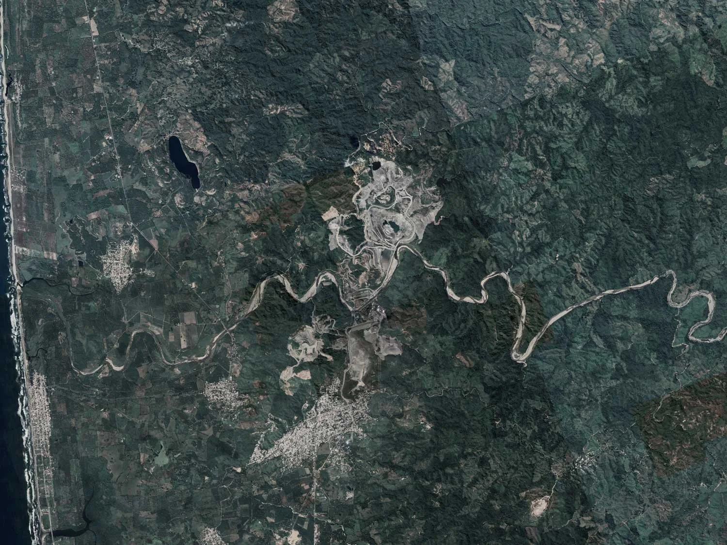 Aerial view of a landscape featuring a winding river through forested and mountainous terrain, with a small town near the coast on the left side of the image.