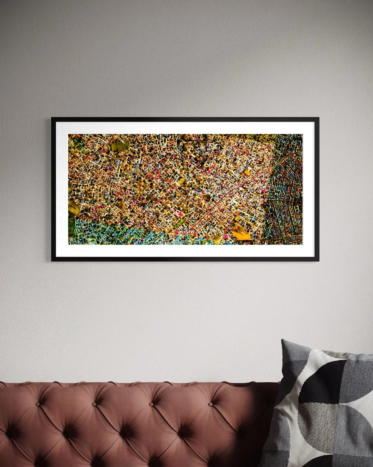A framed aerial map of a city hanging on a white wall above a brown tufted sofa with a black and white patterned pillow.