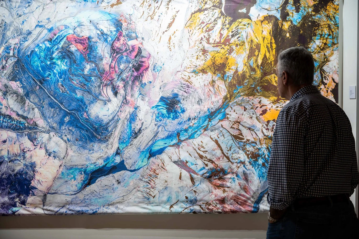 An elderly man with silver hair wearing a black and white checkered shirt admiring an abstract colorful painting in an art gallery. The painting features vivid blue, pink, white, yellow, and brown swirling patterns.