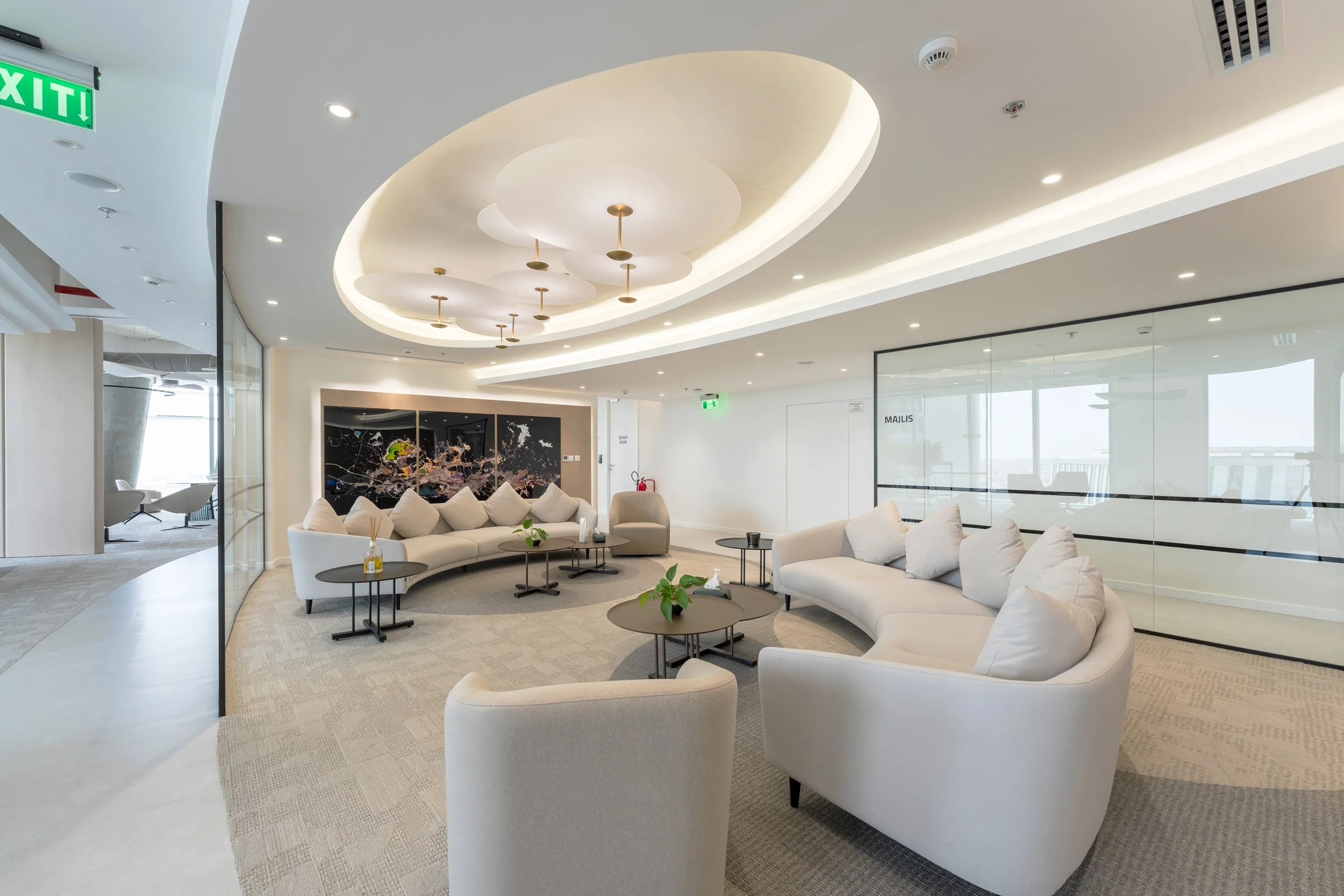 Modern hotel lobby with curved white sofas, black coffee tables, and a large decorative wall art piece. Ceiling has modern white lighting fixtures and a circular recessed design. Large windows with a view outside. Green exit sign and fire extinguisher in the background.